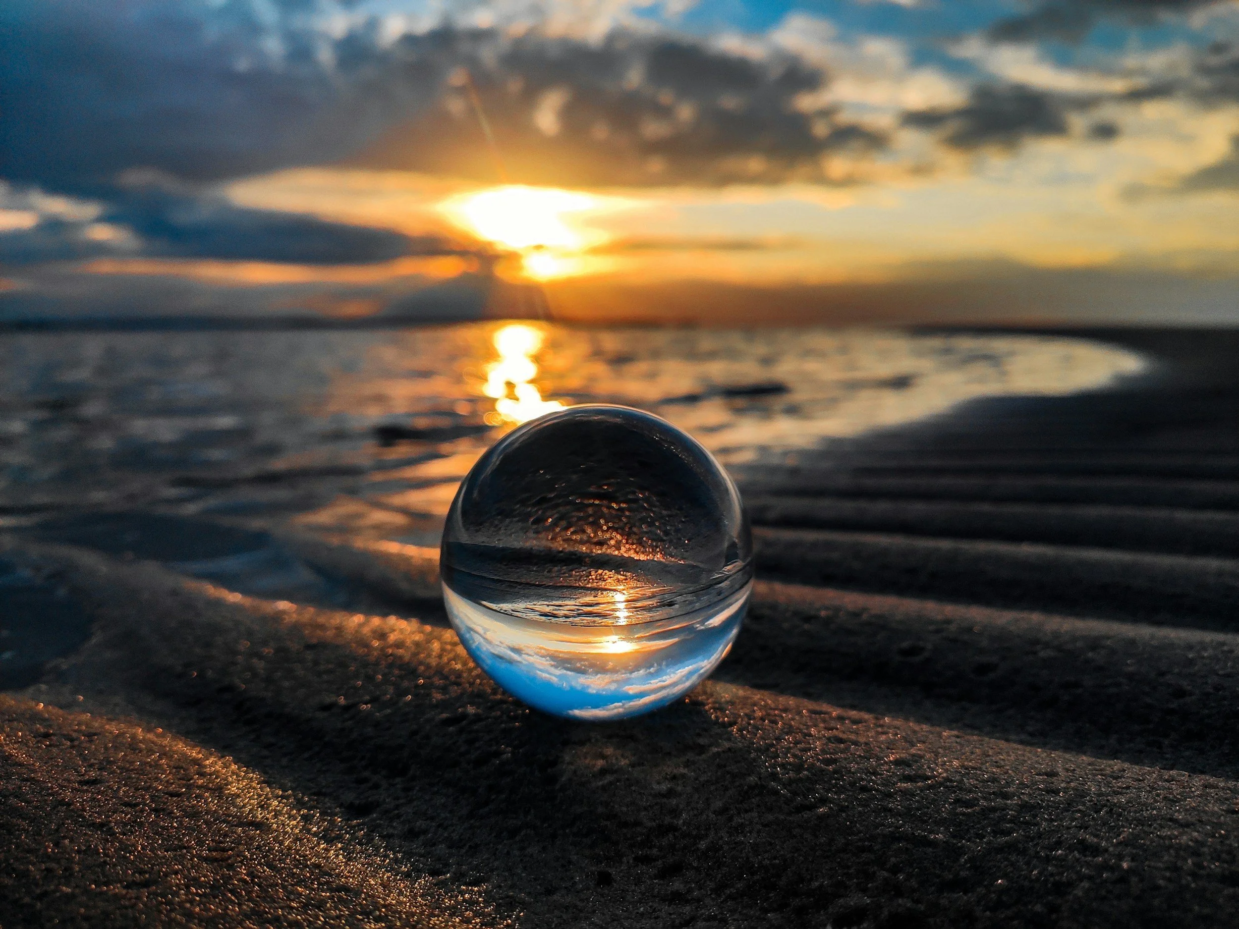 A glass sphere on a sandy beach during sunset, reflecting the sky and water, with clouds and the sun near the horizon.