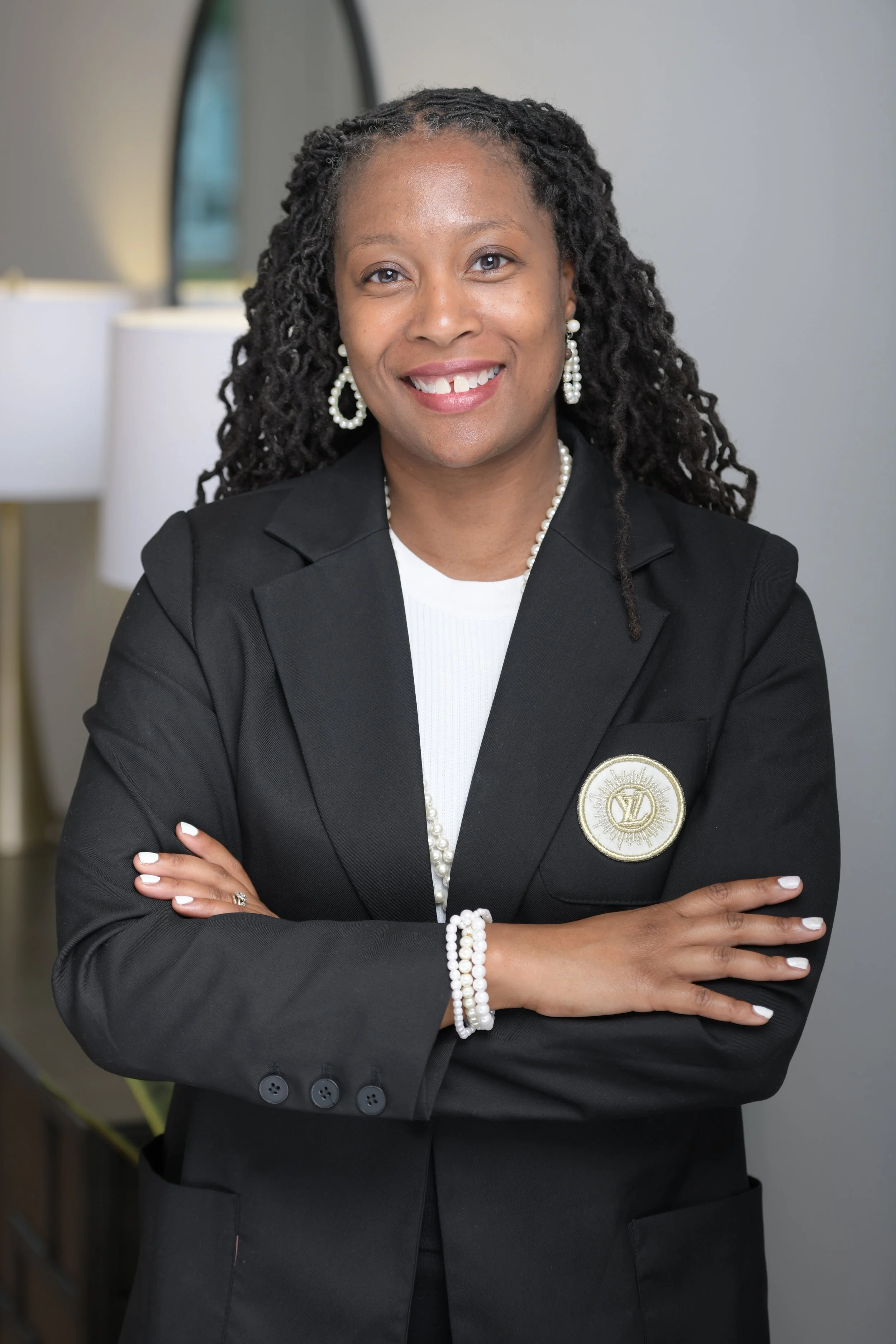 A professional woman smiling with crossed arms, wearing a black blazer with a circular gold badge, pearl jewelry, and earrings, standing in an indoor setting with a lamp and mirror in the background.