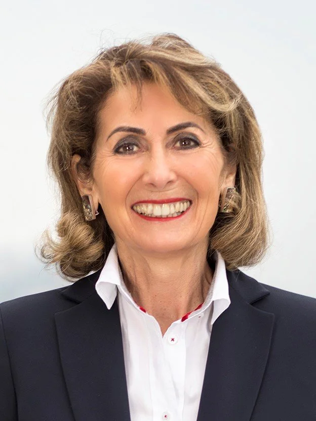 Porträt einer lachenden älteren Frau mit braunen, gewellten Haaren, trägt schwarzen Blazer und weiße Bluse, gegen weißen Hintergrund.