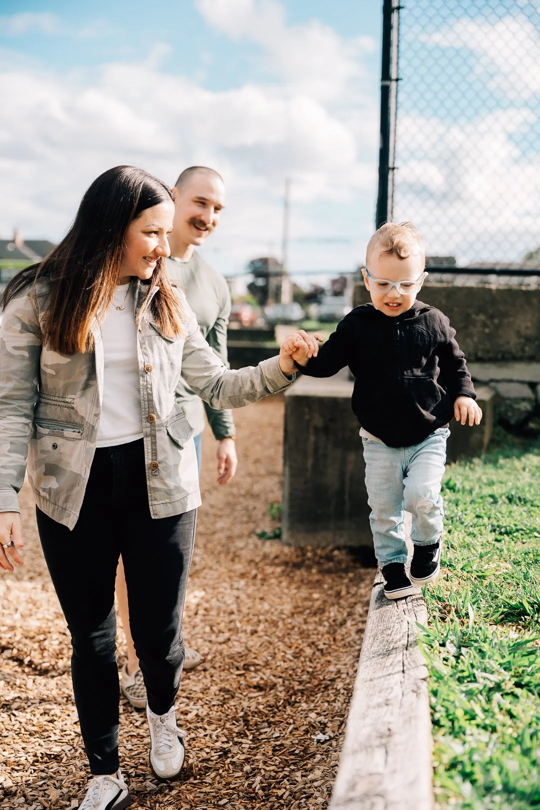 A woman and a man are walking outside with a young boy. The woman is holding the boy's hand as he balances on a wooden beam near a garden bed. The sky is partly cloudy.
