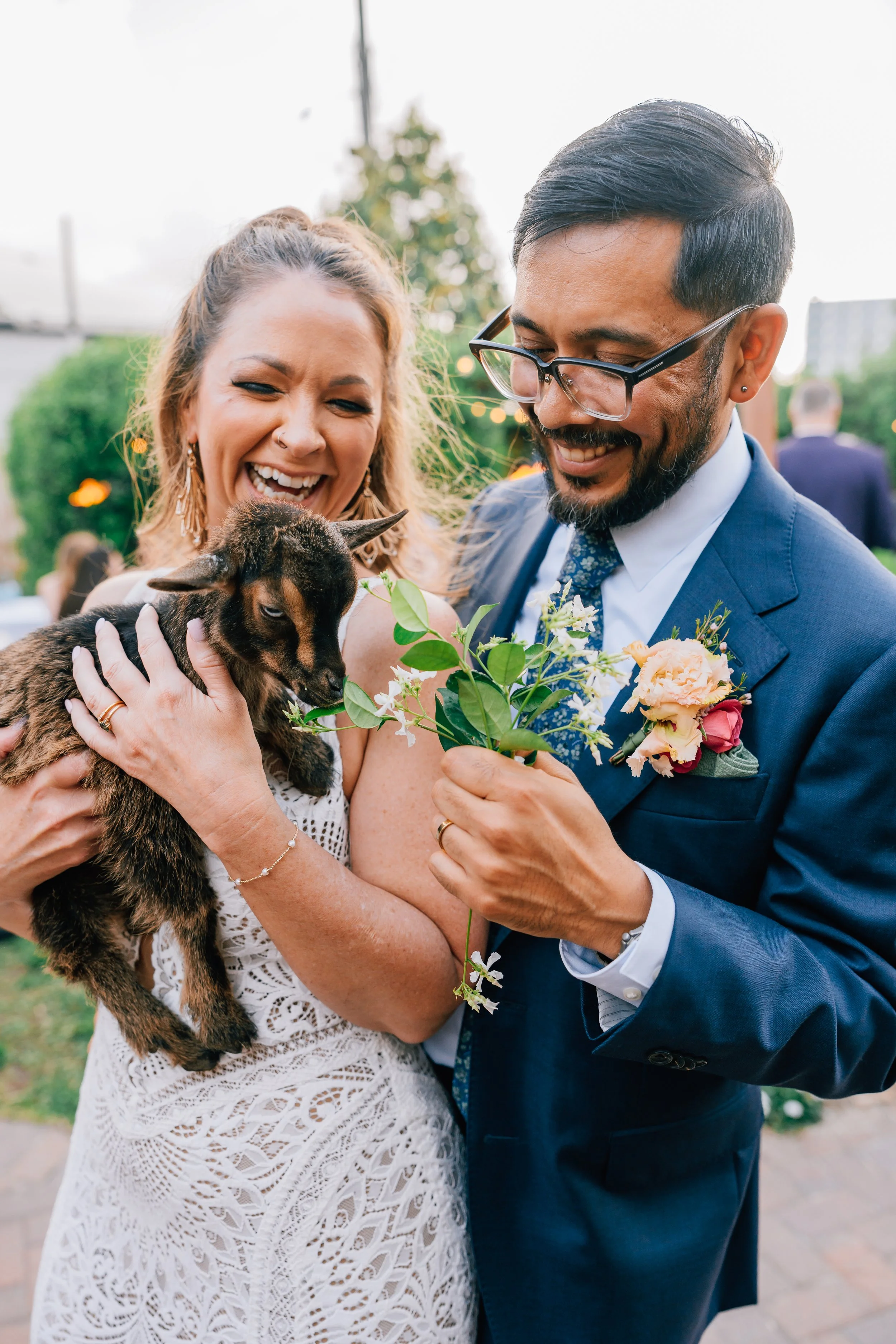 A joyful woman with light skin and wavy blonde hair holding a small brown and black baby goat, standing next to a smiling man with light skin, dark hair, glasses, wearing a blue suit, holding a bouquet of flowers, at their Alaska elopement