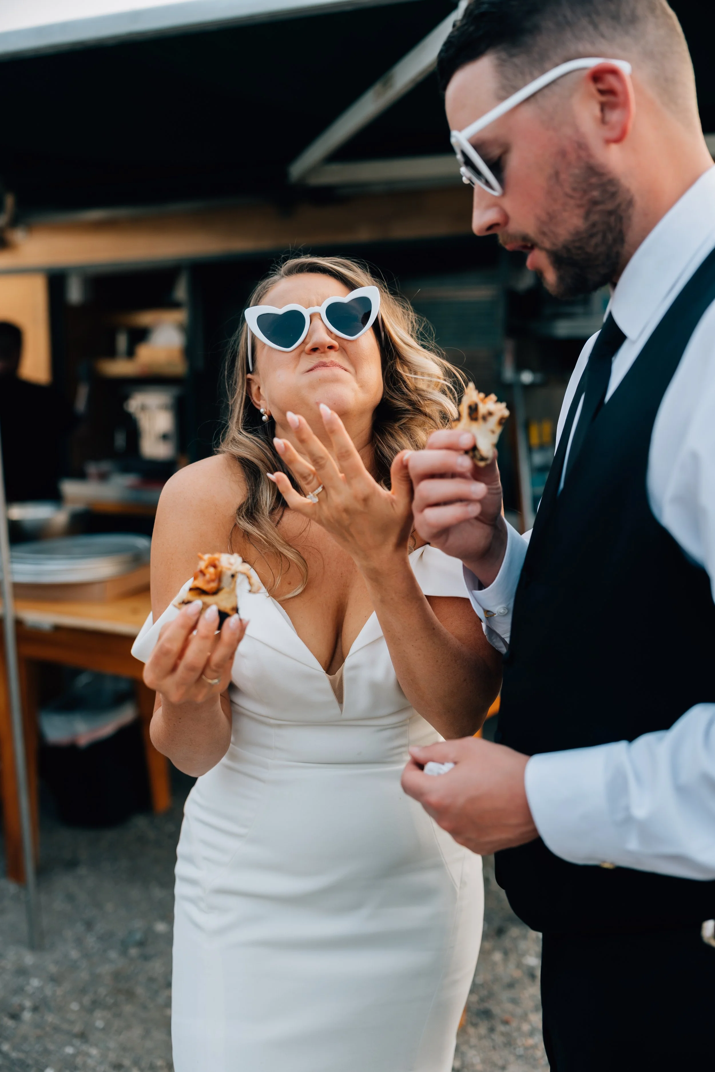 Woman in white dress with heart-shaped sunglasses making a funny face at her Alaska elopement photographer while holding pizza with a man in black vest and white shirt eating pizza