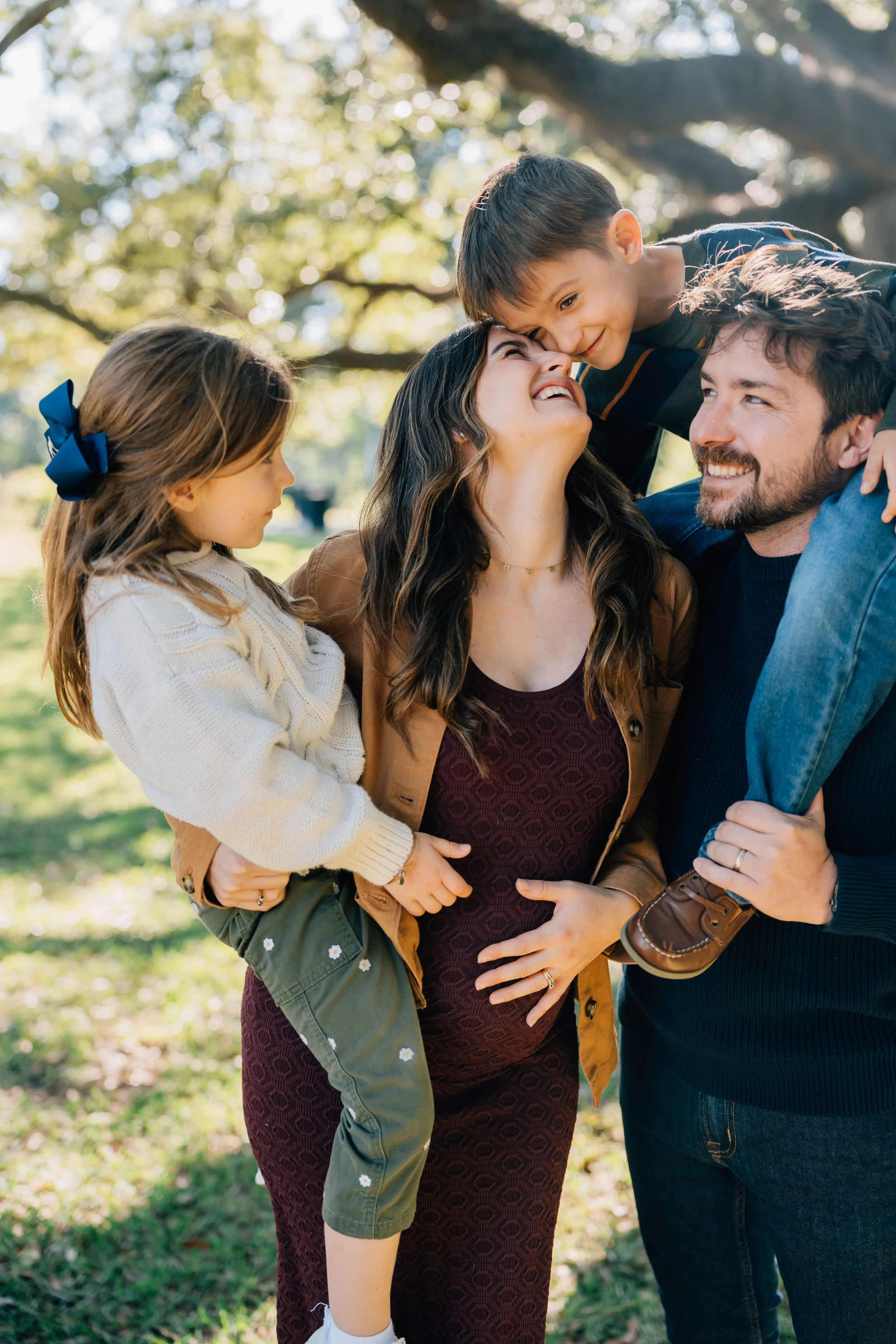 A happy family of four enjoying time together outdoors on a sunny day, with trees in the background. The mother, who is pregnant, is being held by the father. Their son is leaning over her, touching her nose, and the daughter is sitting on her father's arm, looking at her mother.