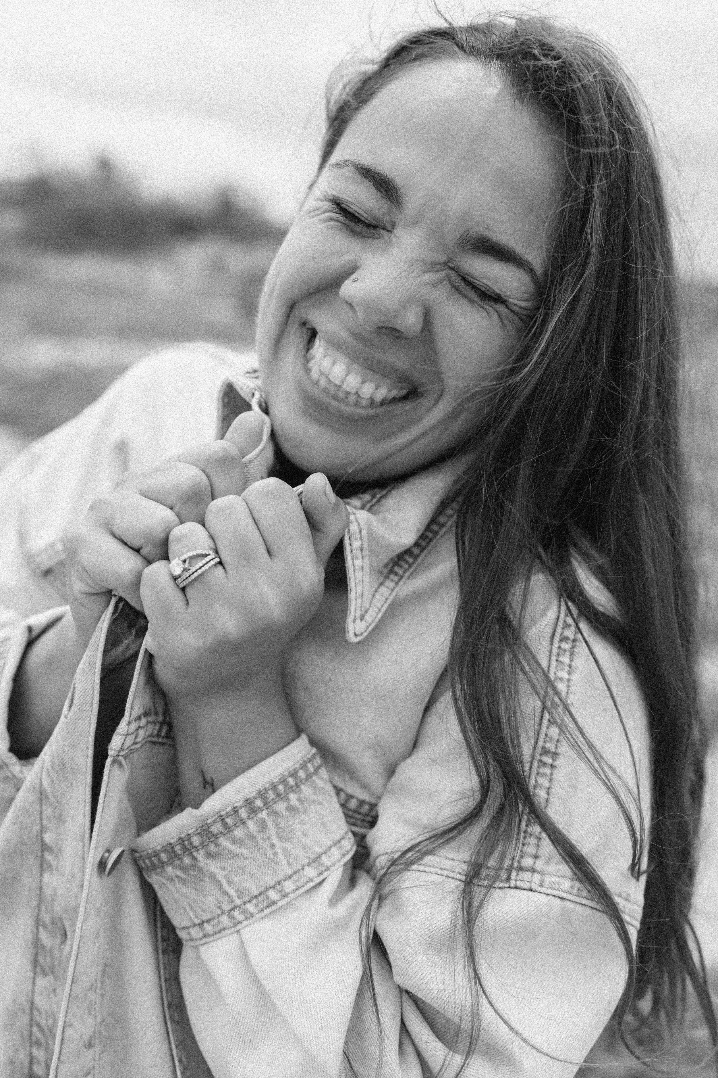 A Juneau Alaska photographer laughing outdoors, with her eyes closed and wearing a denim jacket, holding her neck with her hand and showing off rings.