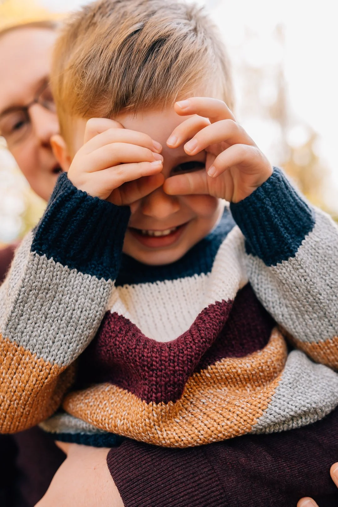 A young boy with blonde hair laughs while covering his eyes with his hands, wearing a multicolored sweater, as an adult holds him and smiles in the background.