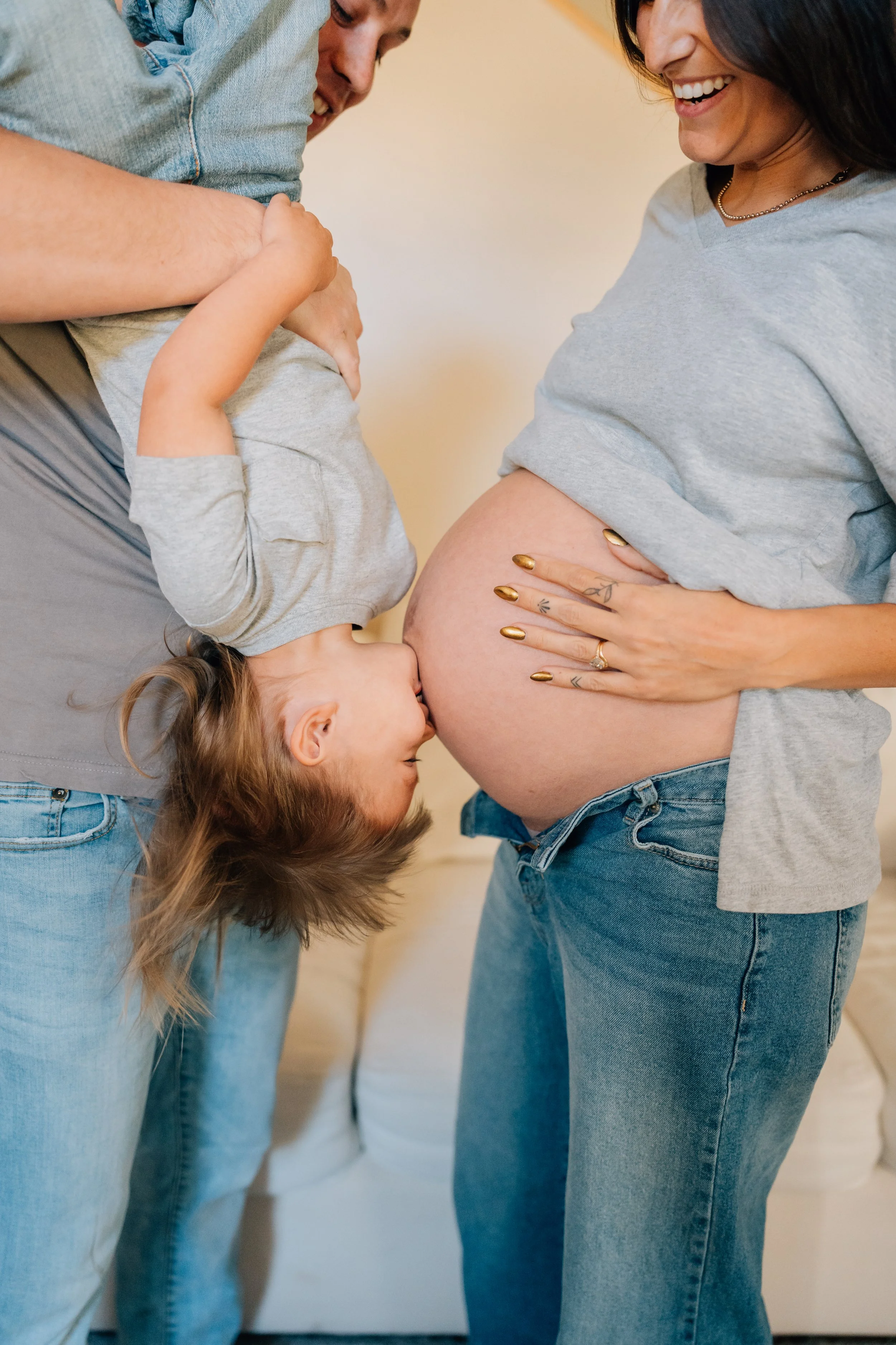 A pregnant woman with blonde hair and tattoos on her fingers is smiling, and a child is kissing her belly while a man holds the child.