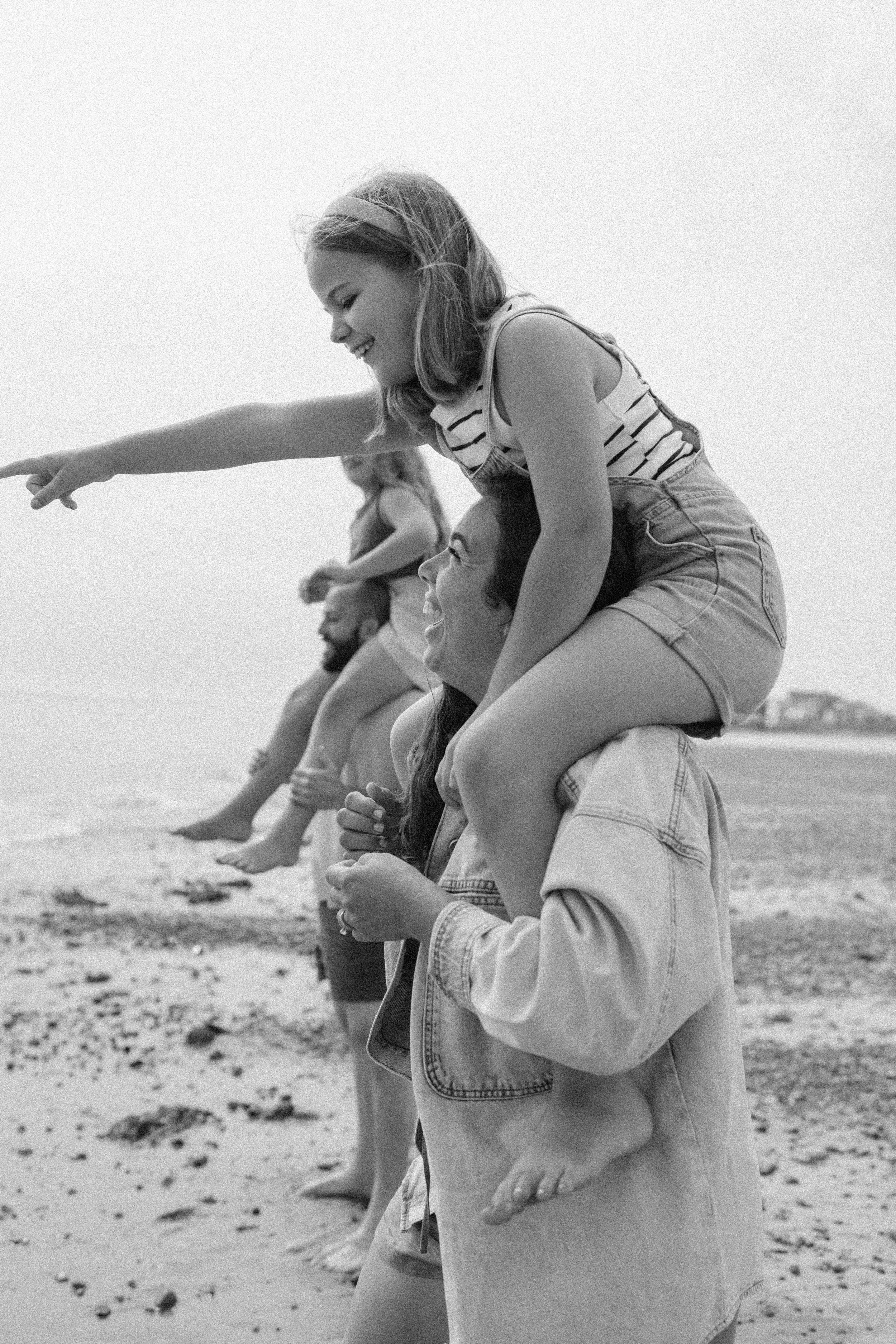 A family of 4 in Juneau Alaska, 3 women and one man, enjoying a beach day. One woman is riding on a man's shoulders, pointing and smiling, while the others stand in line behind them, also smiling and relaxed.