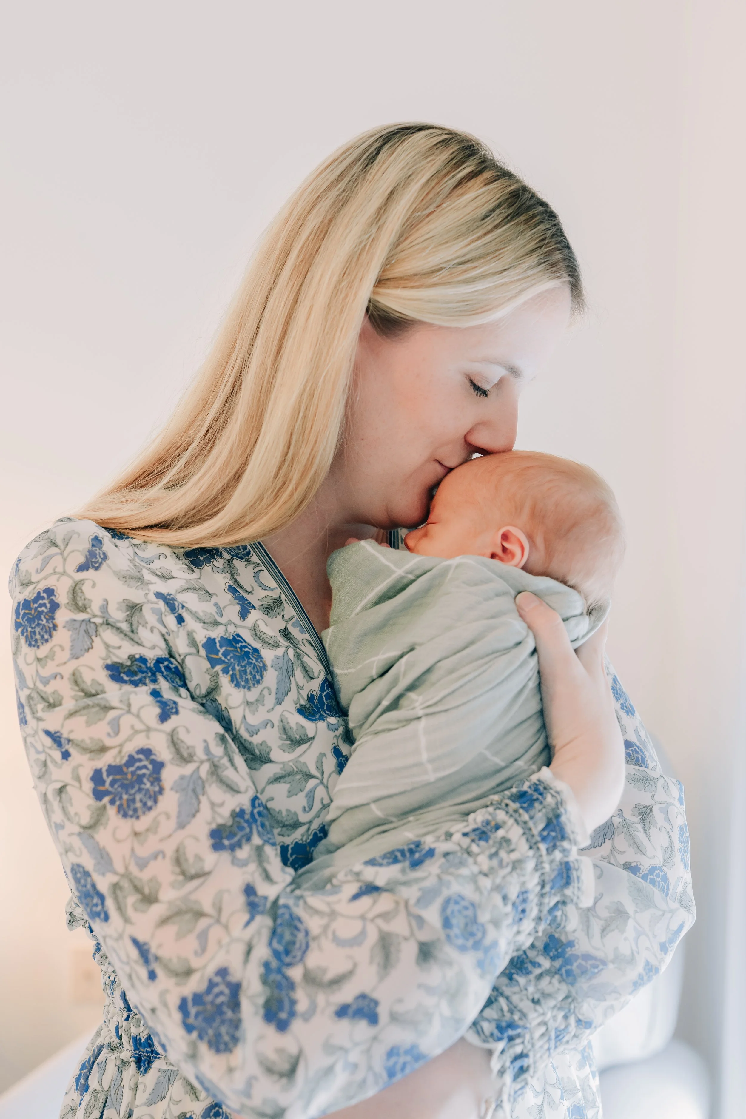 A woman with blonde hair holding a newborn baby close and kissing the baby's forehead in a tender moment for their Juneau newborn photographer