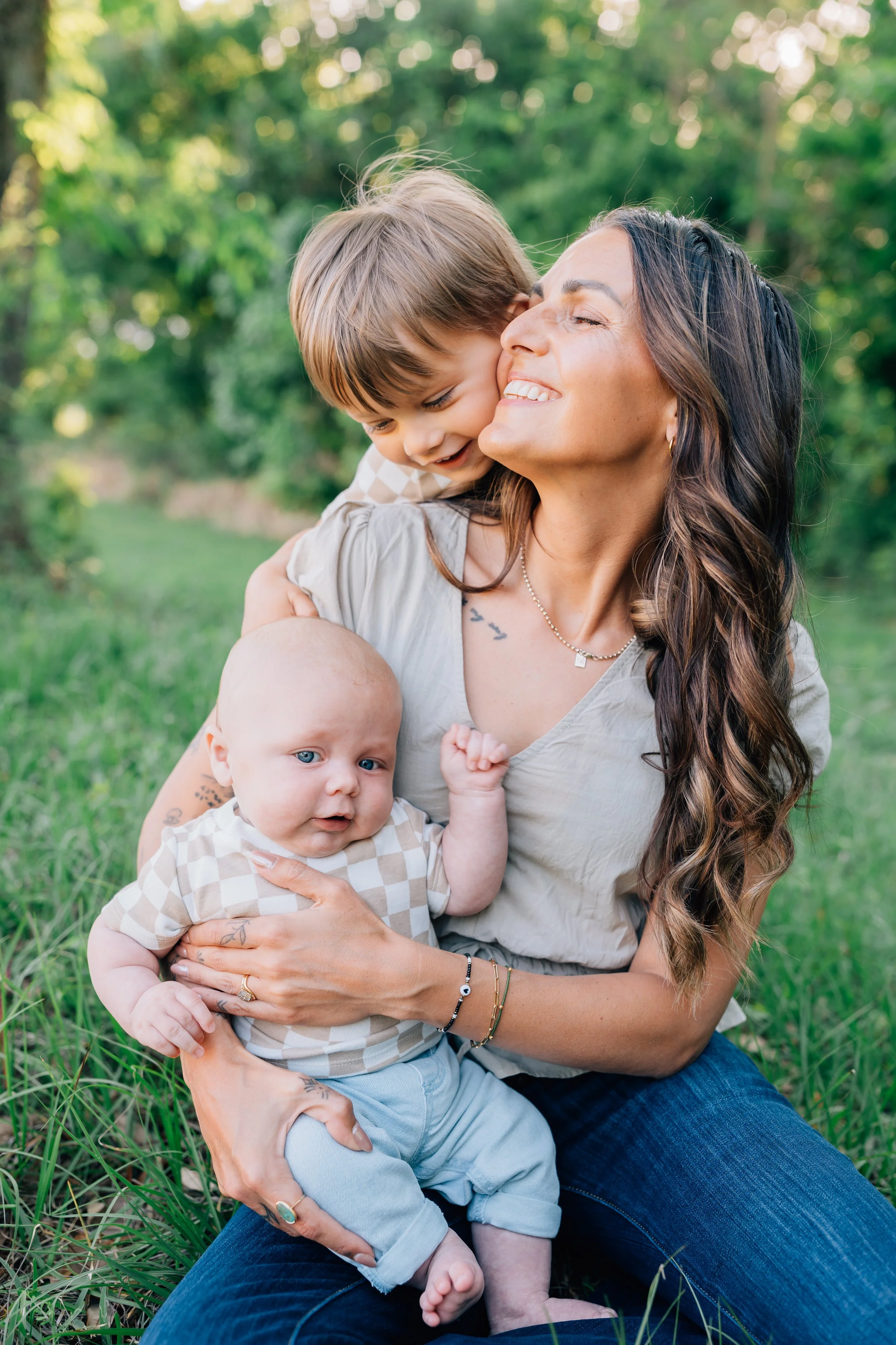 A woman with long brown hair smiling while holding a baby and a young boy outdoors in a grassy area in Juneau Alaska with trees in the background.