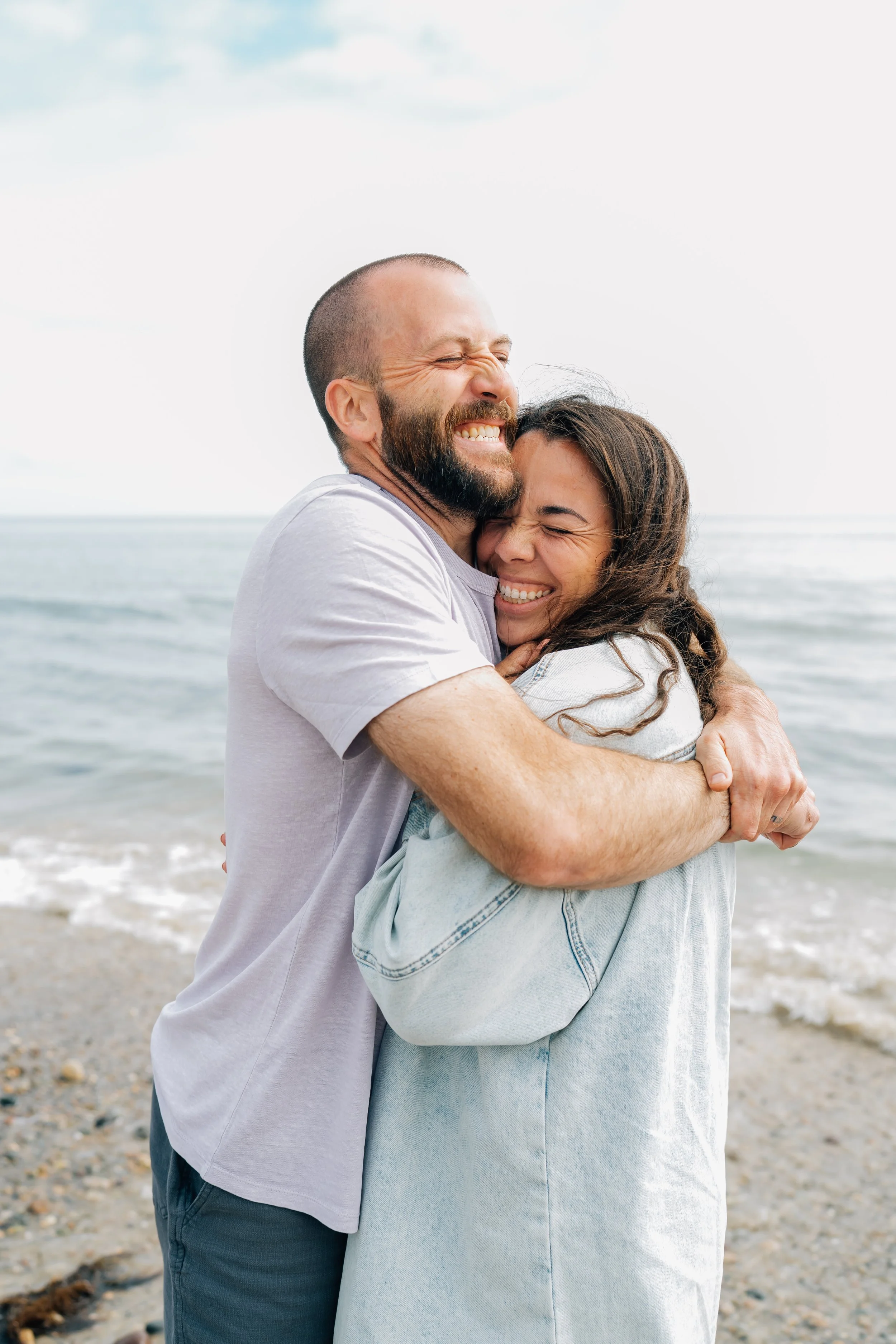 A smiling couple hugging at the beach with the ocean in the background.