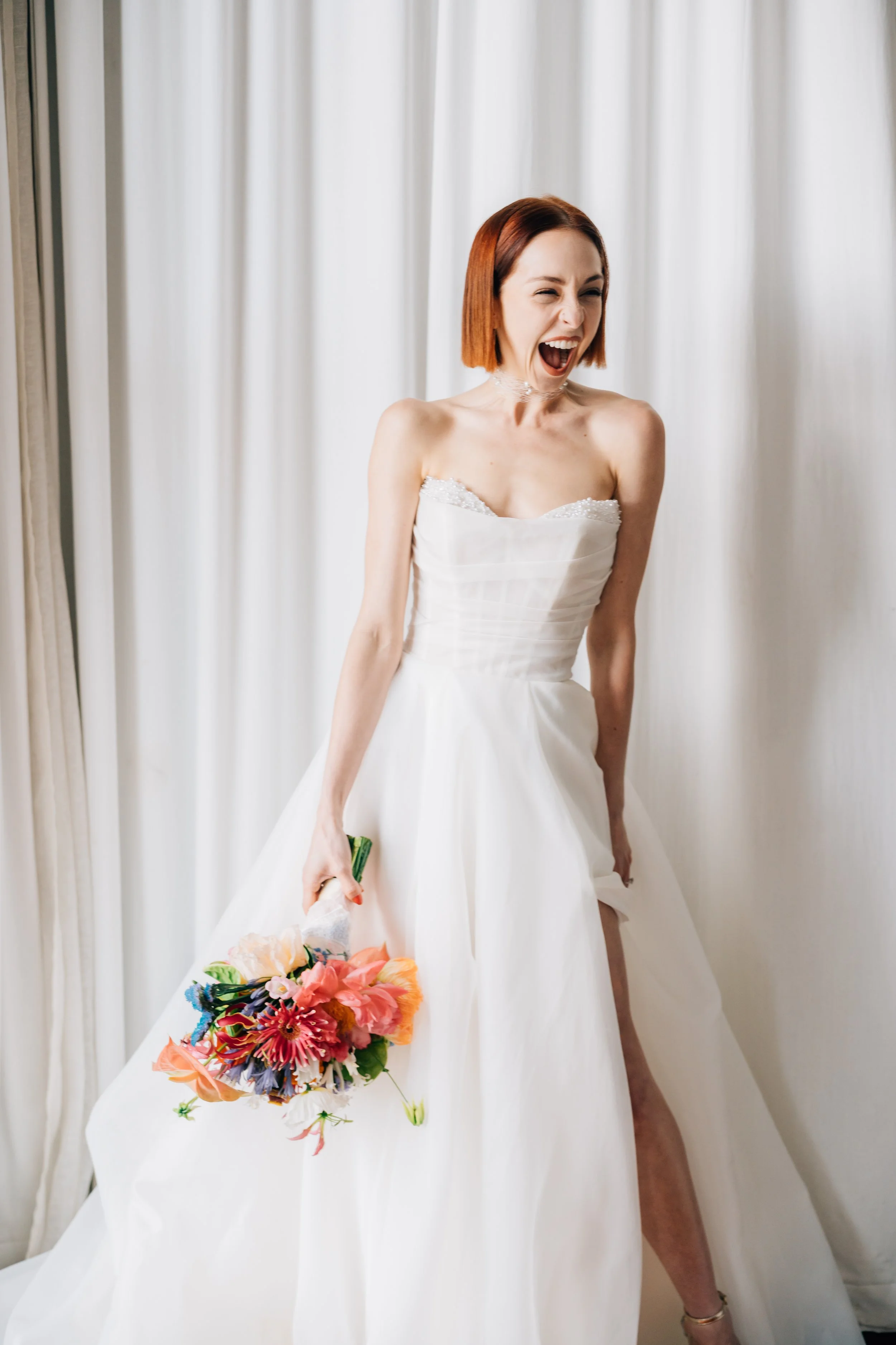 A woman in a white wedding dress laughing and holding a colorful bouquet. She is standing in front of white curtains.