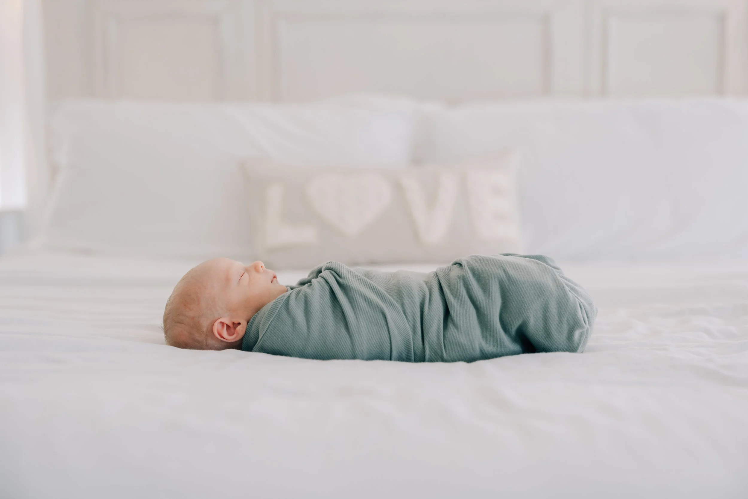 A newborn baby sleeping on a white bed with a decorative pillow that spells out 'LOVE' with a heart symbol at Juneau newborn photos