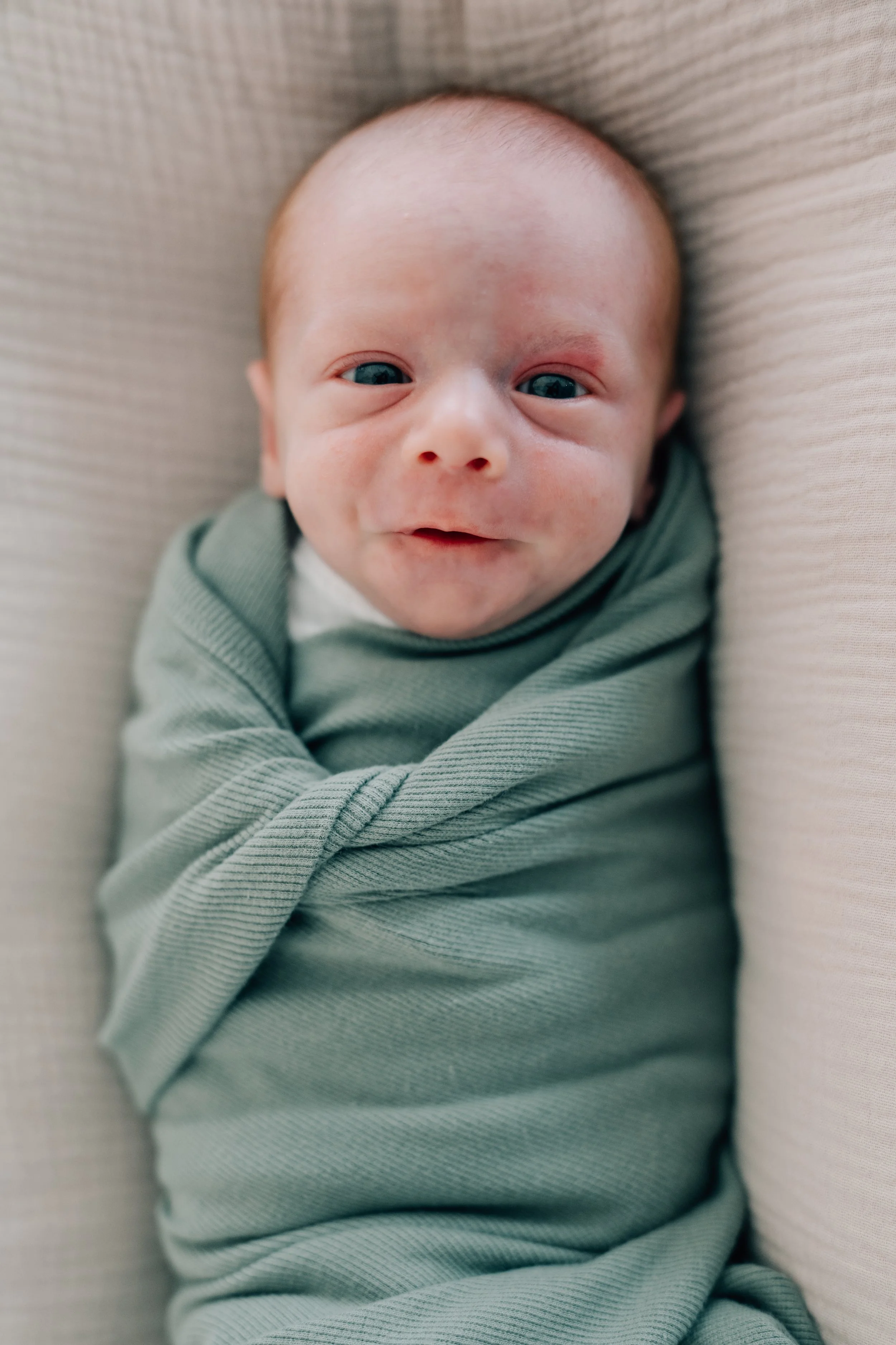 Close-up of a smiling baby wearing a green outfit, lying on a light-colored textured surface.