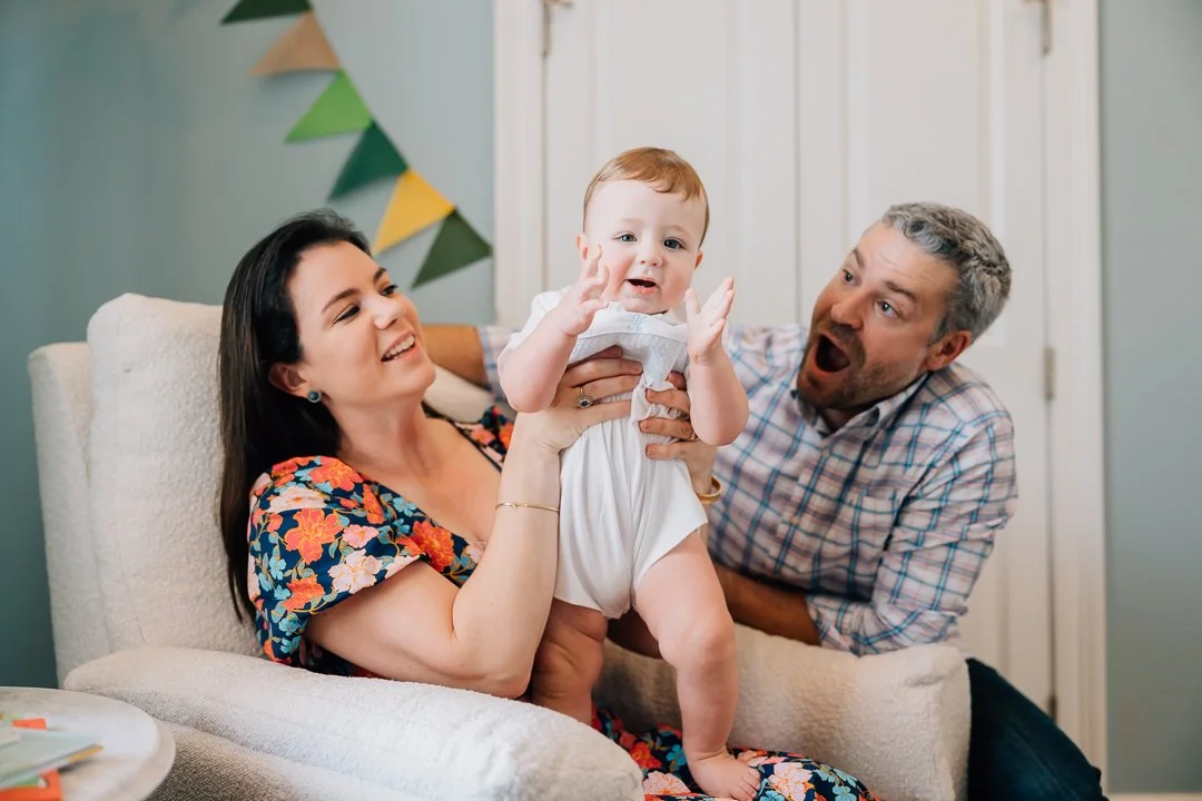 A family of three, a woman, a man, and a baby, enjoying a playful moment together in a living room. The woman is holding the baby, who has a surprised expression, while the man is reacting with excitement.