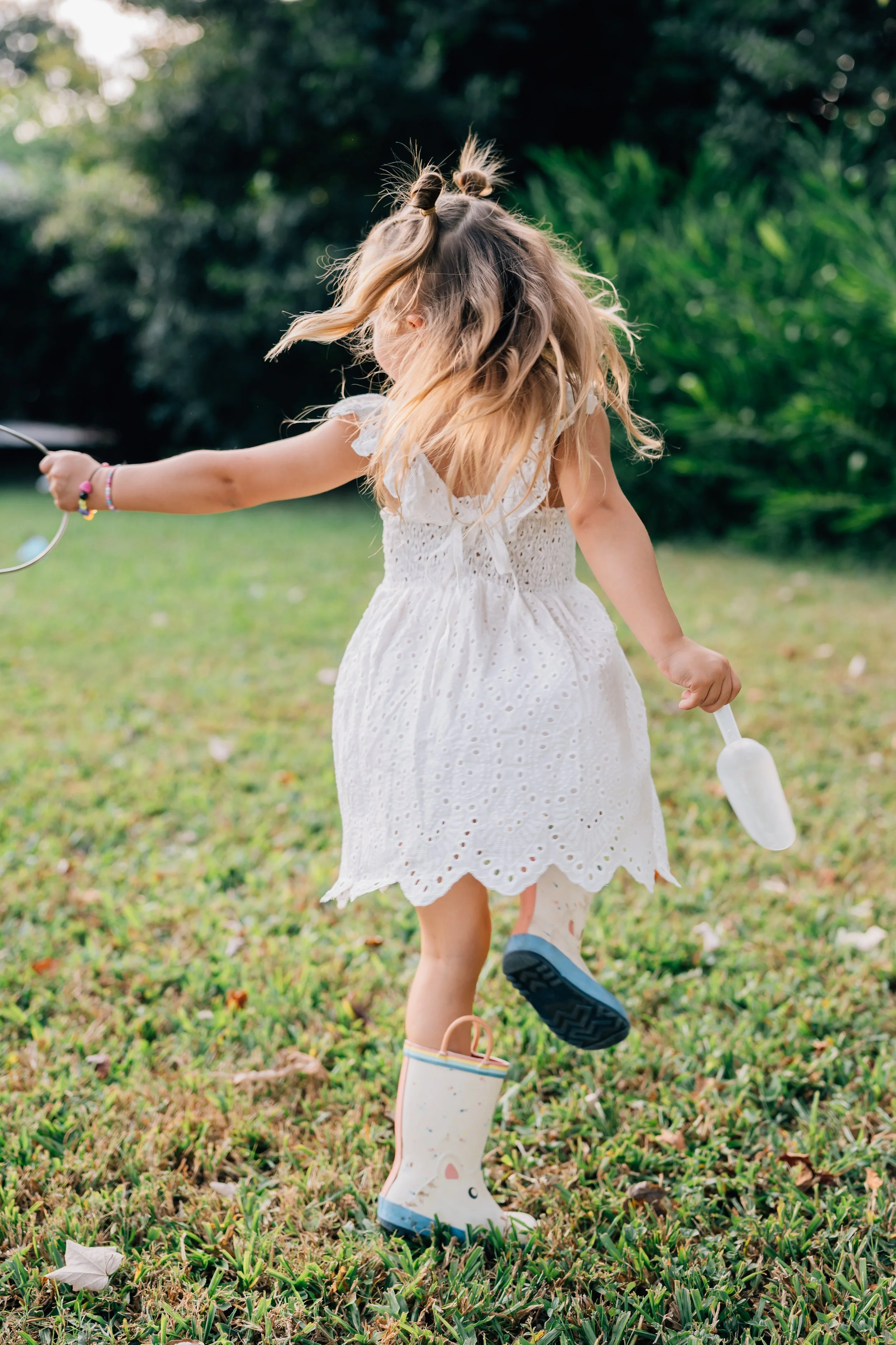 Young girl in a white dress playing outdoors, wearing rain boots, holding a small shovel, with wind blowing her hair.