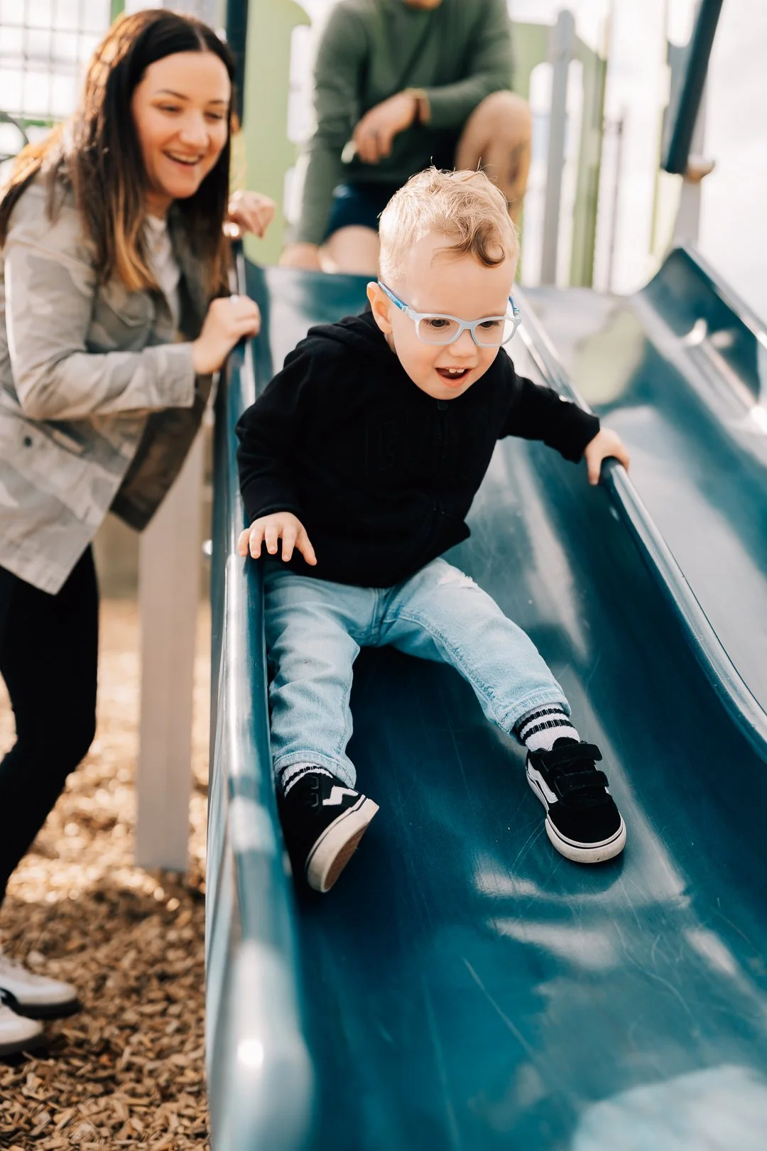 A young boy with glasses sliding down a playground slide, smiling, with a woman nearby cheering him on.