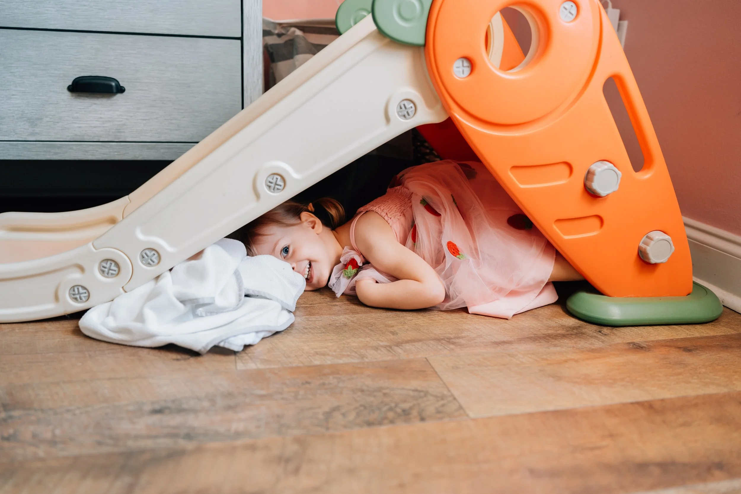 A young girl with brown hair in pigtails, wearing a pink dress with strawberry embroidery, is lying underneath a tipped-over orange and cream plastic slide on a wooden floor, smiling and resting her head on a white cloth.