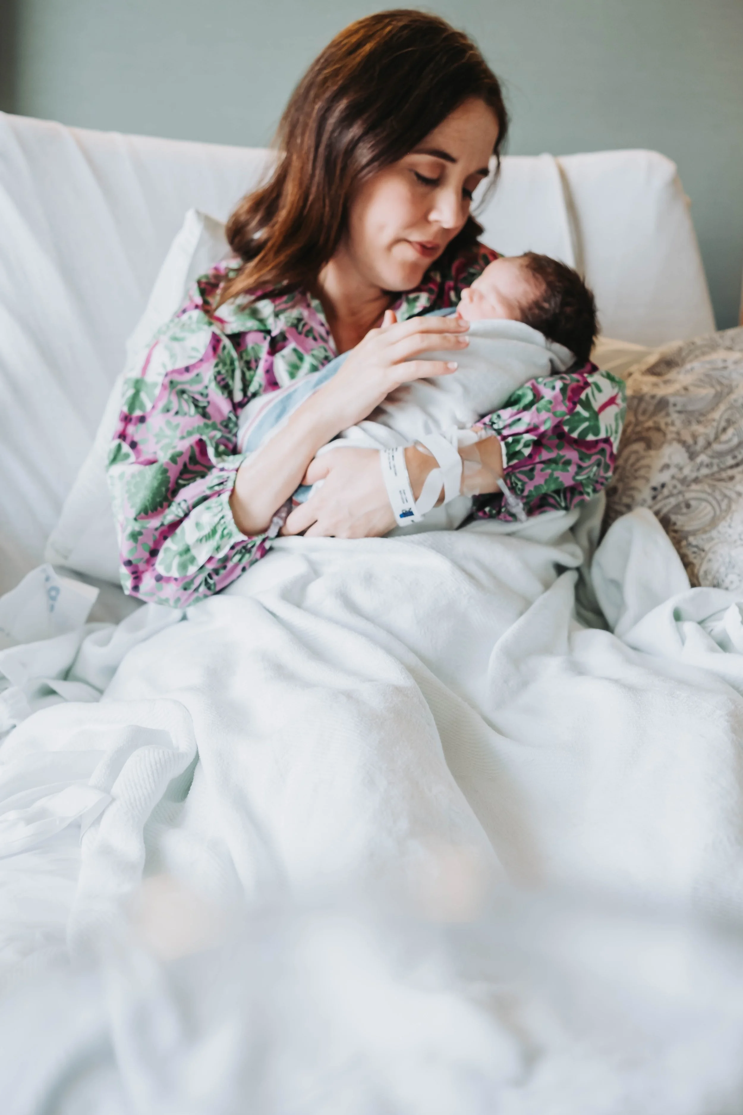 A woman in a floral hospital gown holds a newborn baby in a white blanket, sitting on a hospital bed in Juneau Alaska fresh 48 photos