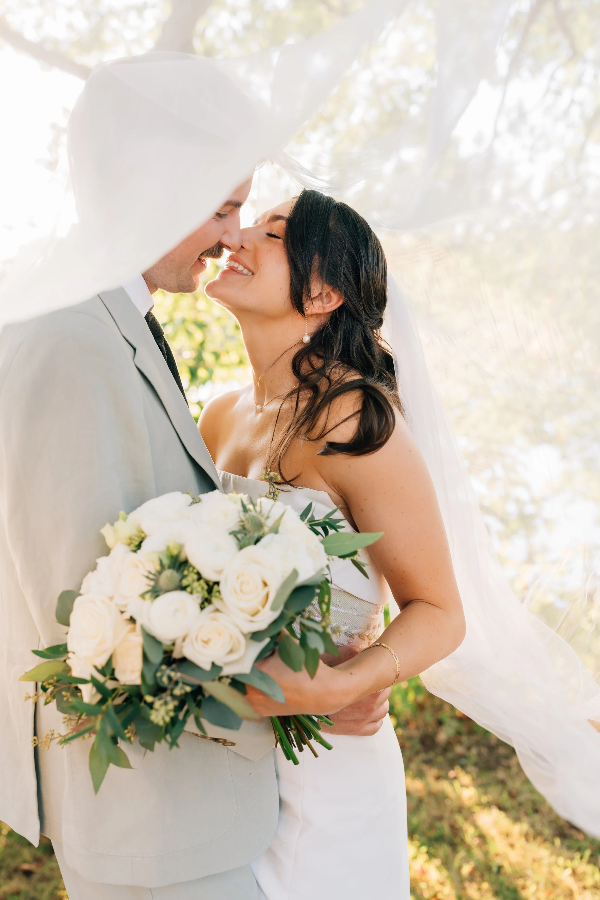 A bride and groom are closely embracing at their Alaska elopement, smiling with their noses touching. The bride is holding a bouquet of white roses and greenery, and her veil is flowing in the breeze. 