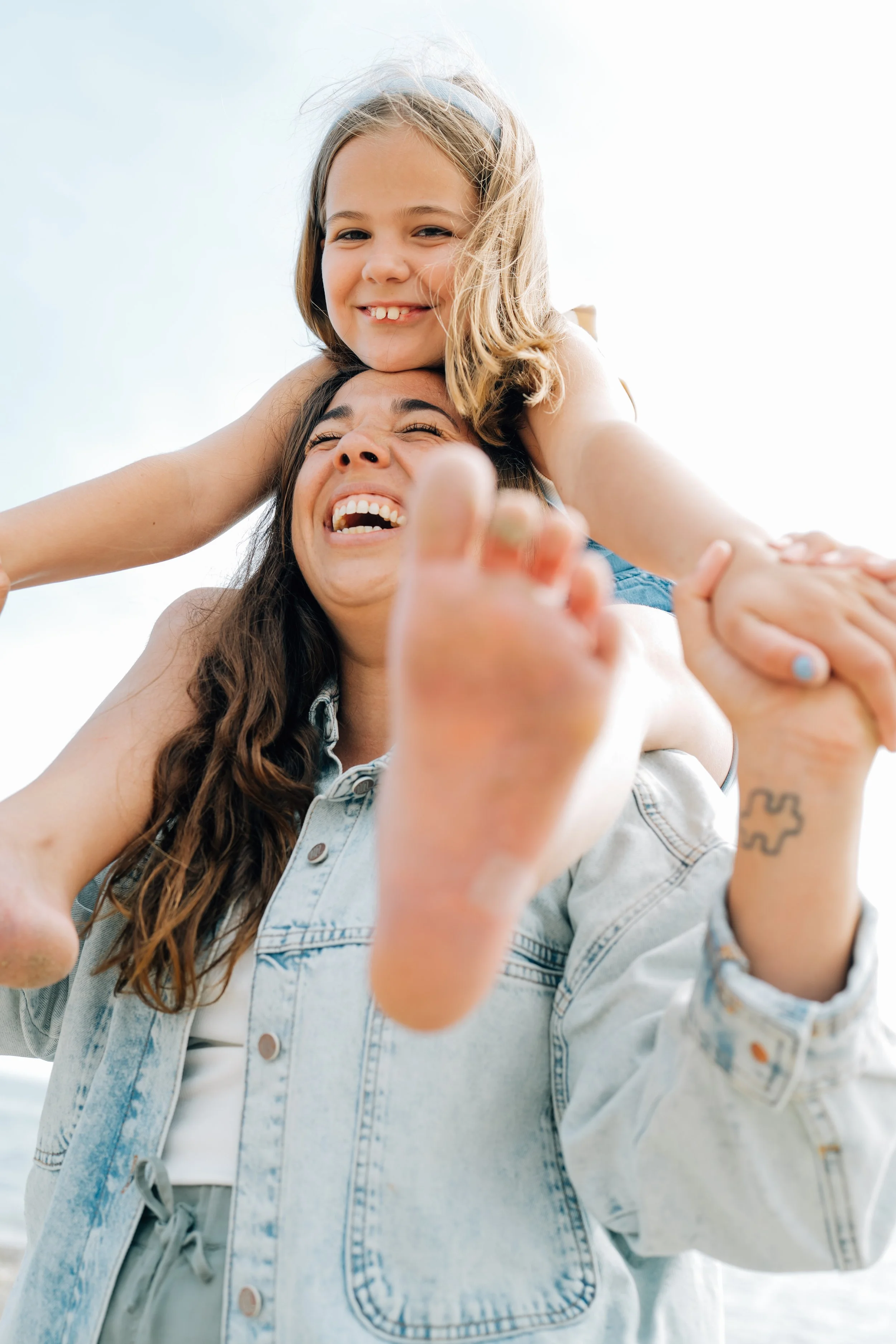 A woman with long brown hair carrying a young girl with blonde hair on her shoulders, both smiling and enjoying a moment together outdoors.