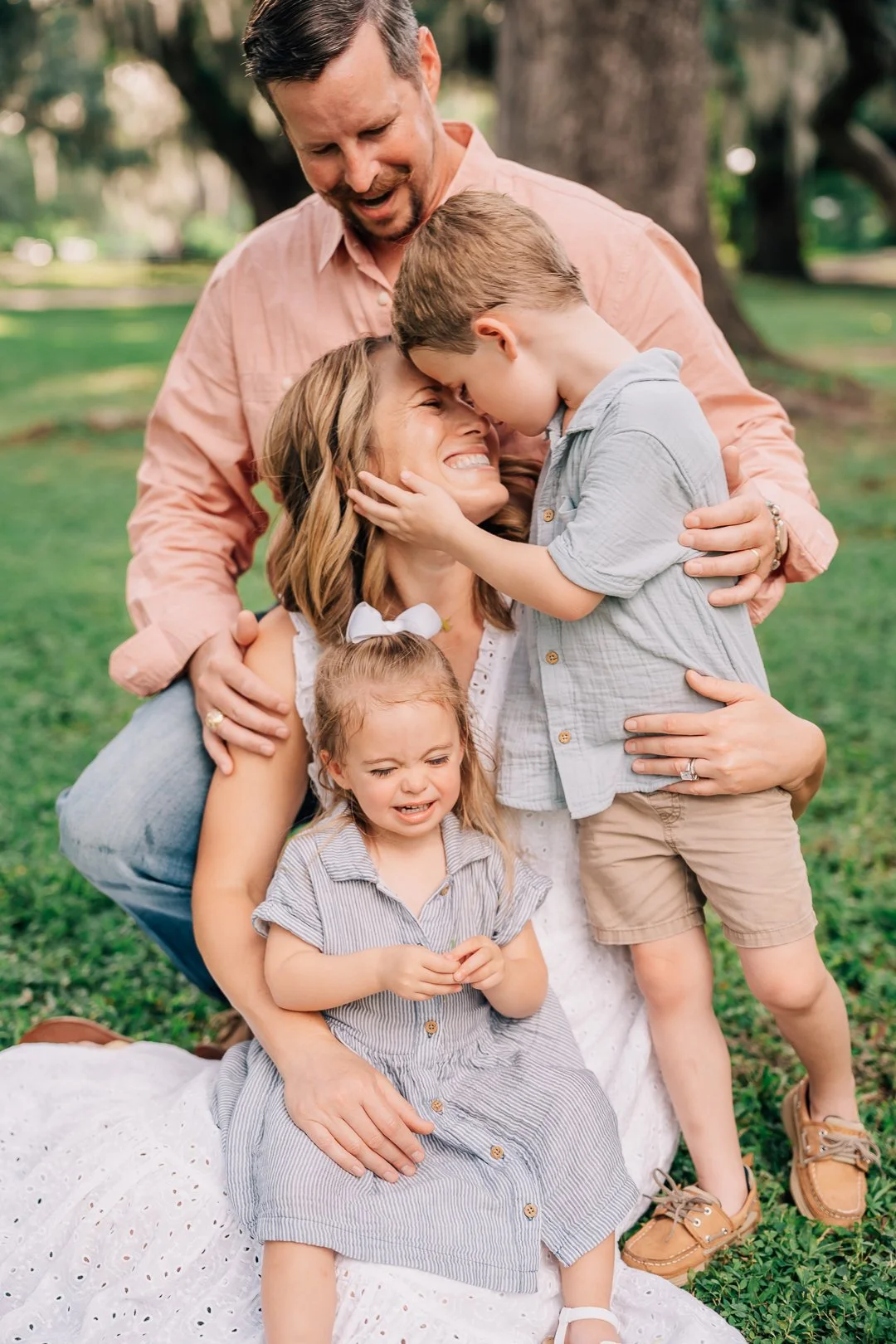 A family of five outdoors on green grass with trees in the background, embracing and smiling, with two young girls and a man and woman, all appearing happy and loving.