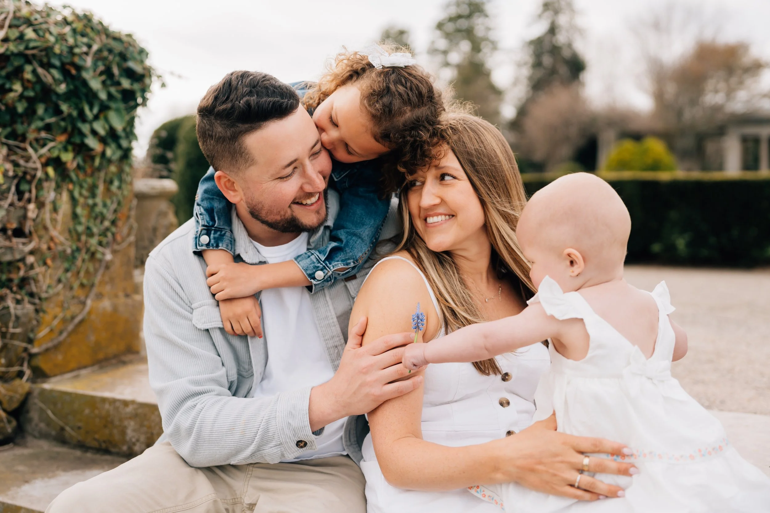 Family outdoors: parents and two children, one girl and one baby girl, enjoying a playful moment, woman holding a blue flower, people smiling and engaging affectionately.