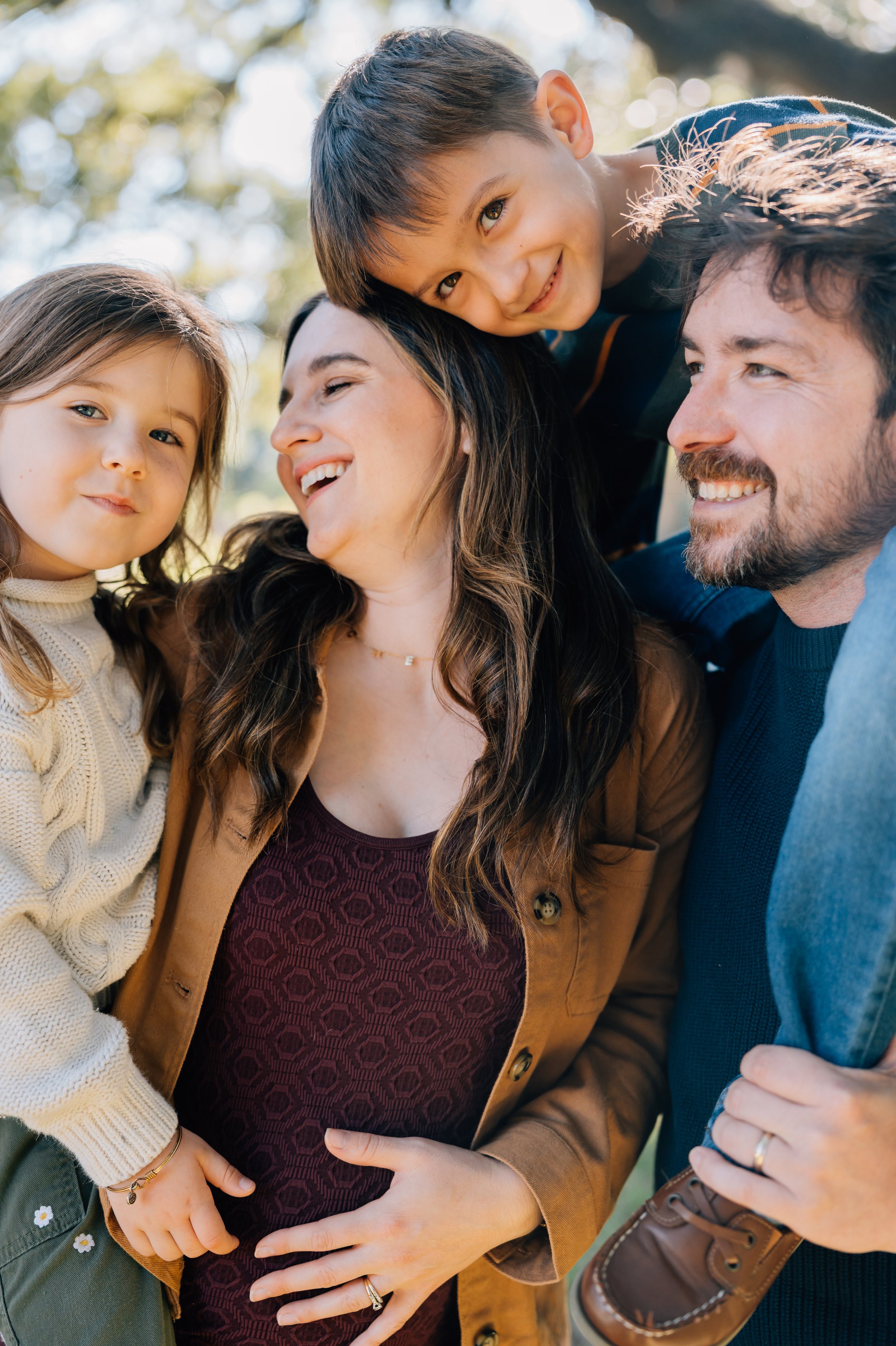 A family of four outdoors enjoying a happy moment together. The woman is holding her pregnant belly, and a young girl and a boy are close to her, with the boy leaning over her shoulder. Everyone is smiling and looking happy.