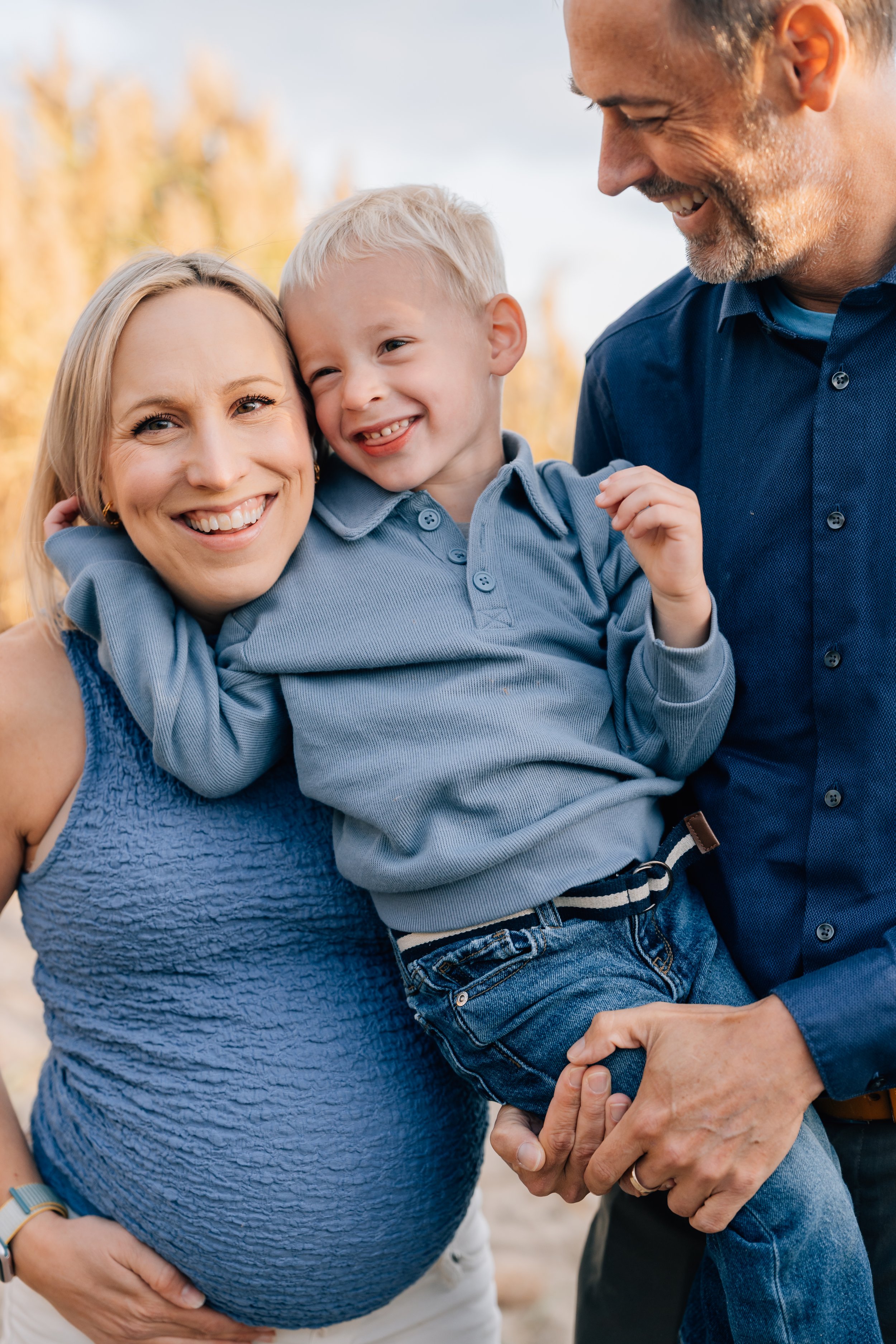 A happy family of three outdoors, with the mother holding a smiling young boy, and the father supporting him, all smiling.