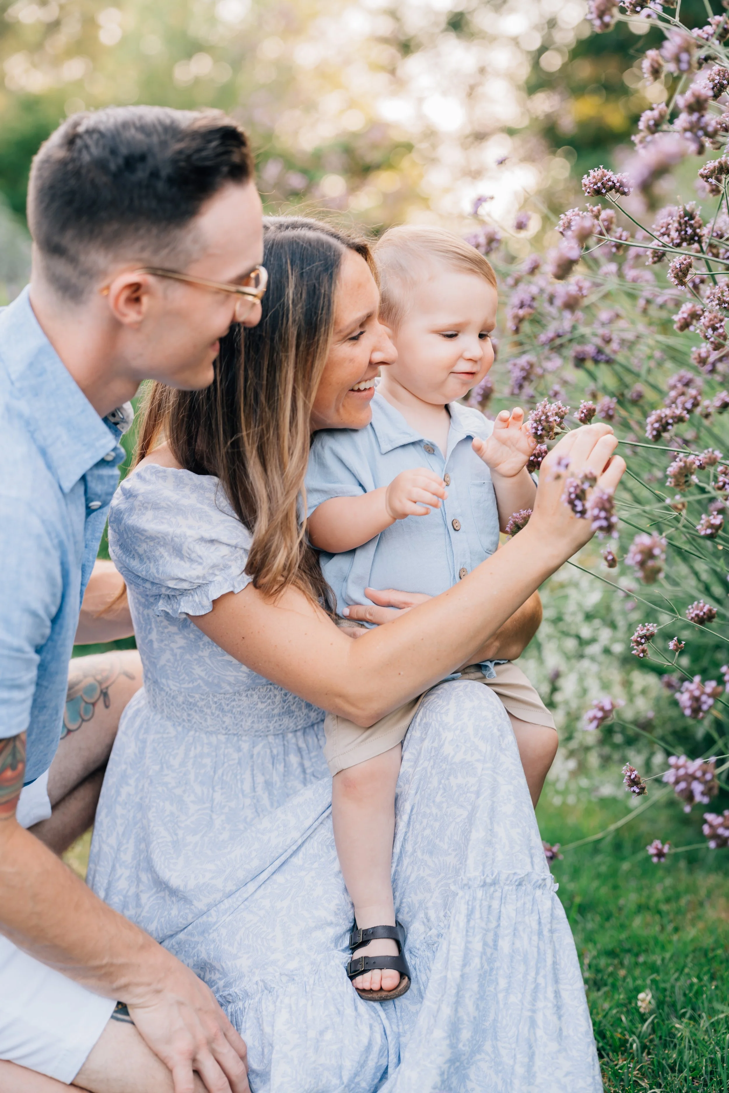 A happy family of four enjoying nature outdoors, sitting together near blooming purple flowers. The mother is holding a young child and touching the flowers, while the father is kneeling beside them, all smiling and looking at the flowers.