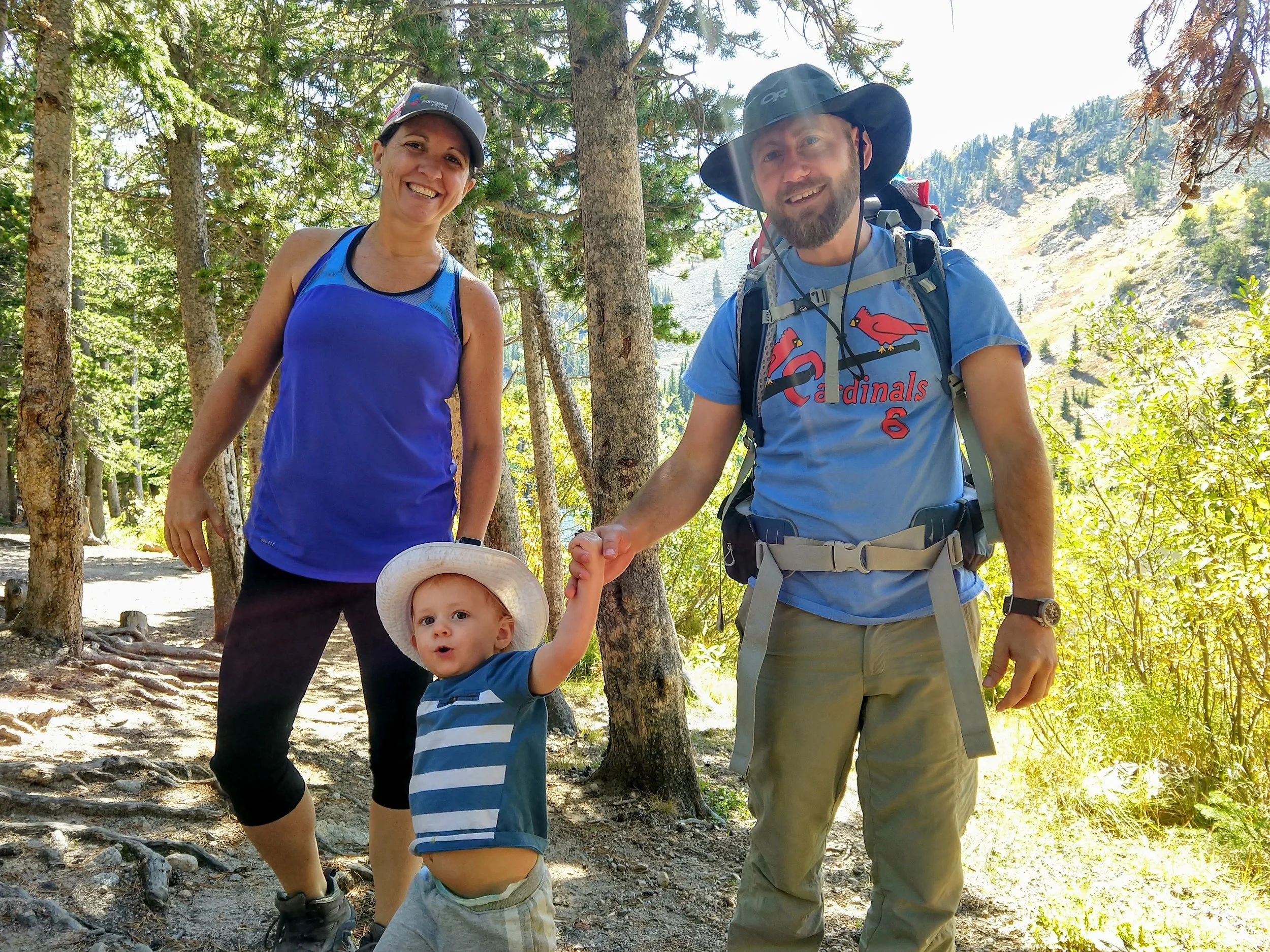 A woman, a man, and a young boy on a hiking trail in a forest. The woman is smiling and wearing a blue sleeveless top and a baseball cap. The man, wearing a wide-brimmed hat and a blue t-shirt with a red bird logo, is holding hands with the young boy. The boy is looking at the camera with a surprised expression, wearing a striped t-shirt and a hat. Trees and mountain scenery are visible in the background.