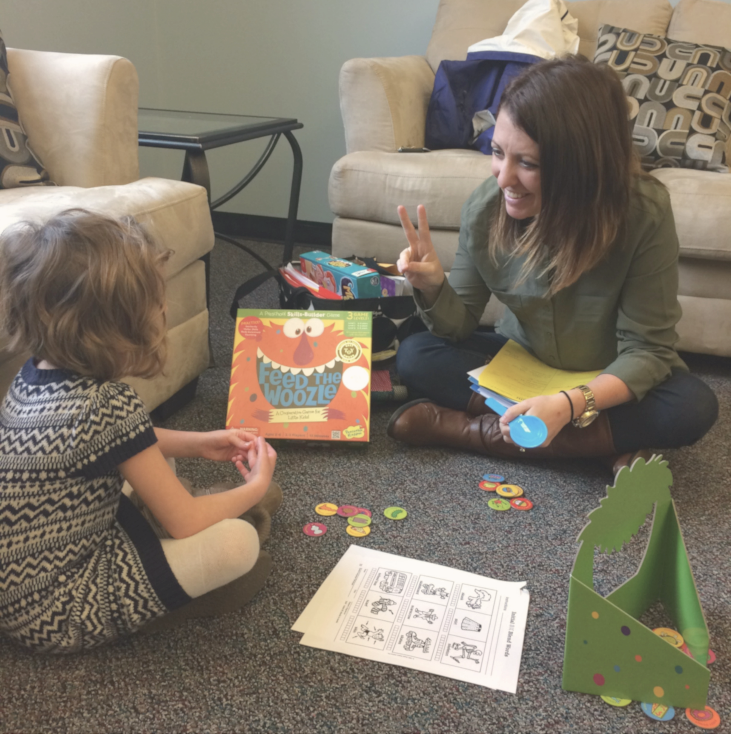 A woman smiling providing speech therapy services while sitting on the floor facing a young child. They are surrounded by various children's activities and educational materials.