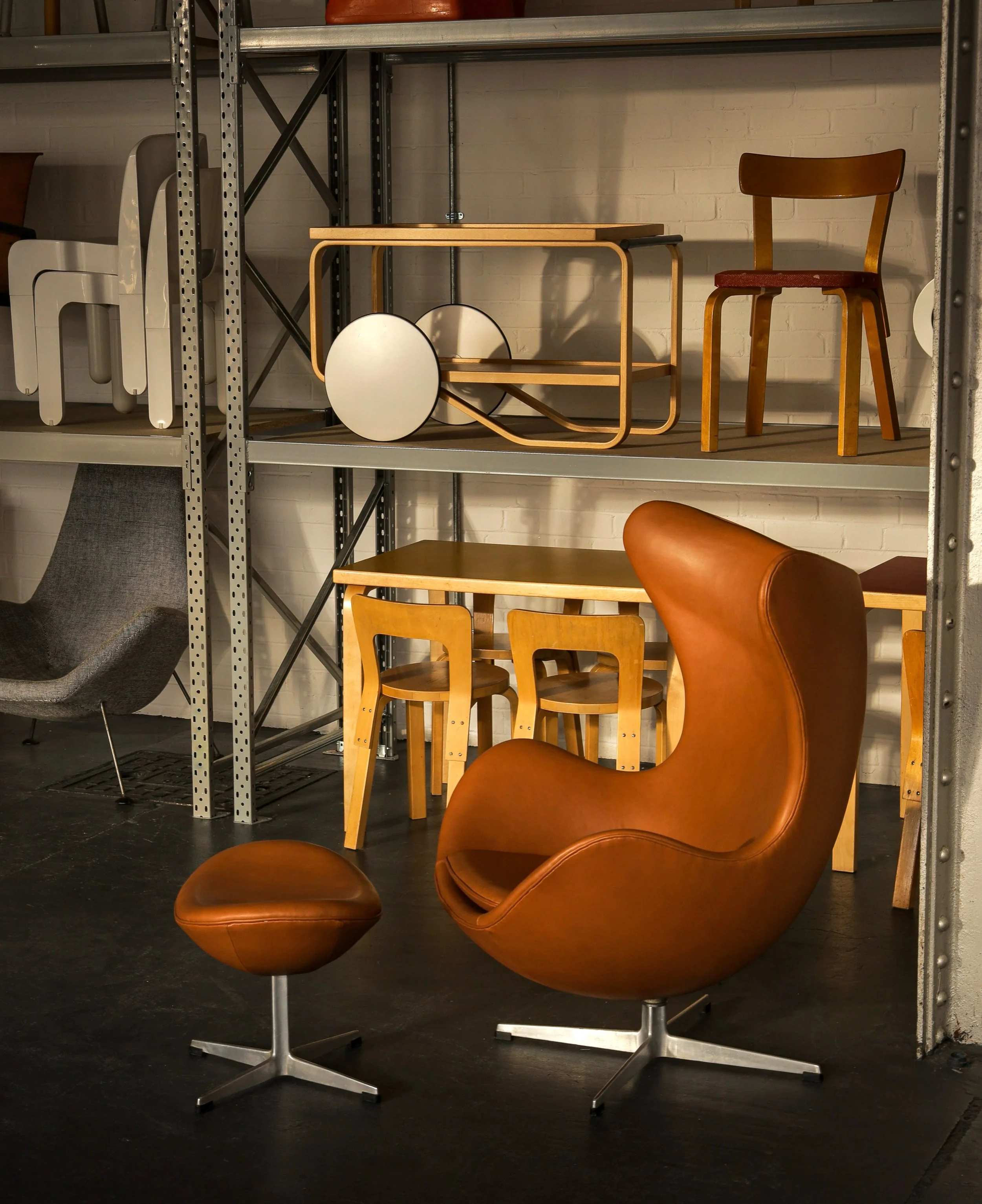Collection of various iconic Finnish design chairs and stools arranged in a warehouse with metal shelving and a brick wall.Featuring Alvar Aalto, Arne Jacobsen, Jean Prouve, Vico Magistretti
