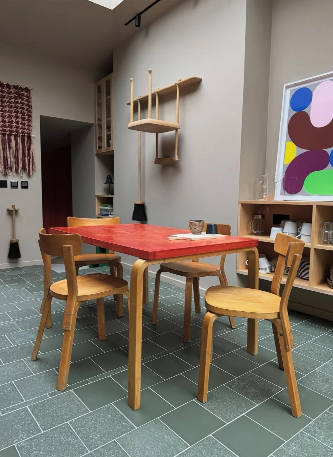A dining area with a red table and four wooden chairs, a wall-mounted shelf, wall decor, and a cabinet with dishes and cups on green tiled flooring.