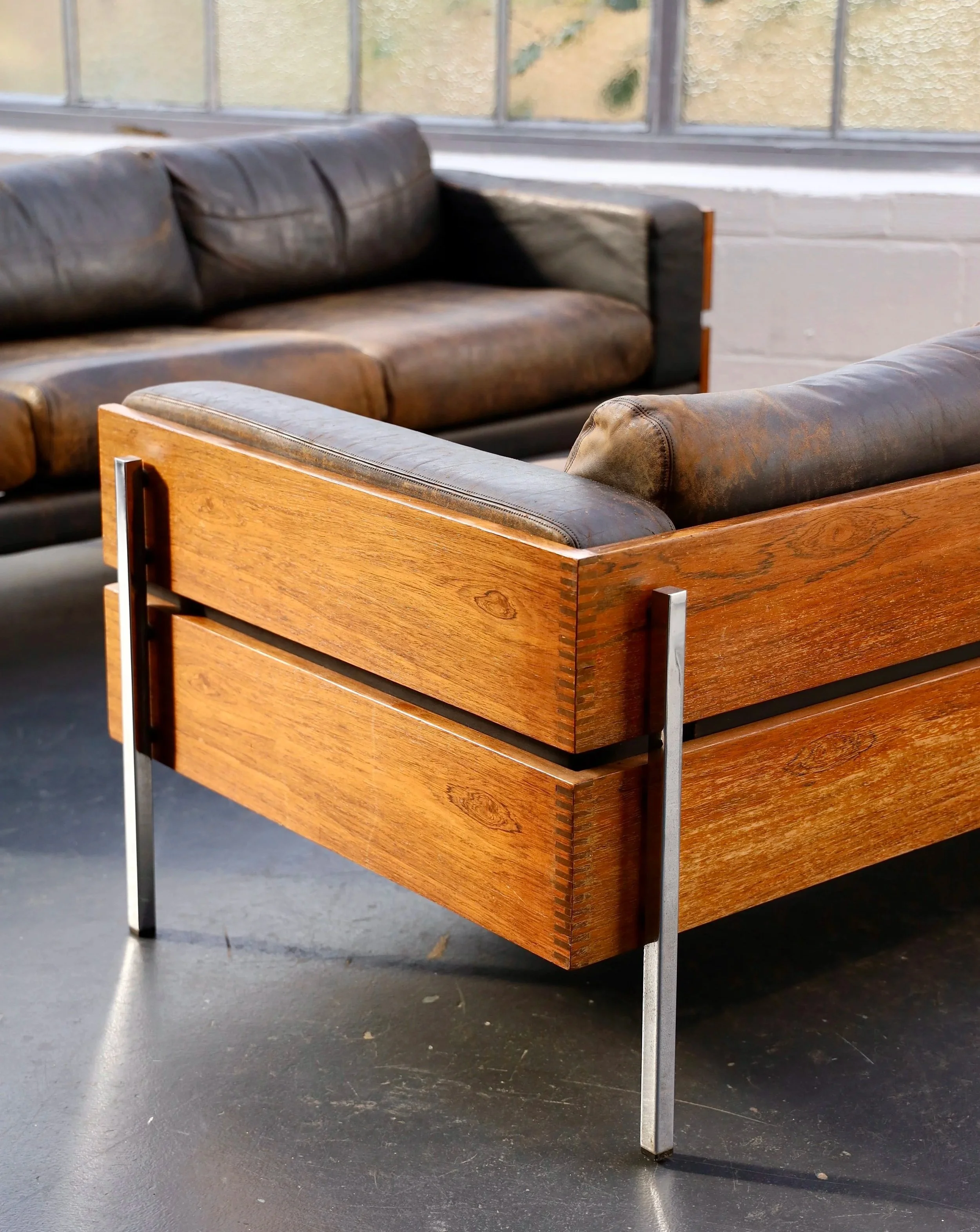 Two vintage Robin Day forum sofas leather couches with metal and wood frame in an indoor setting near large windows with a textured glass.