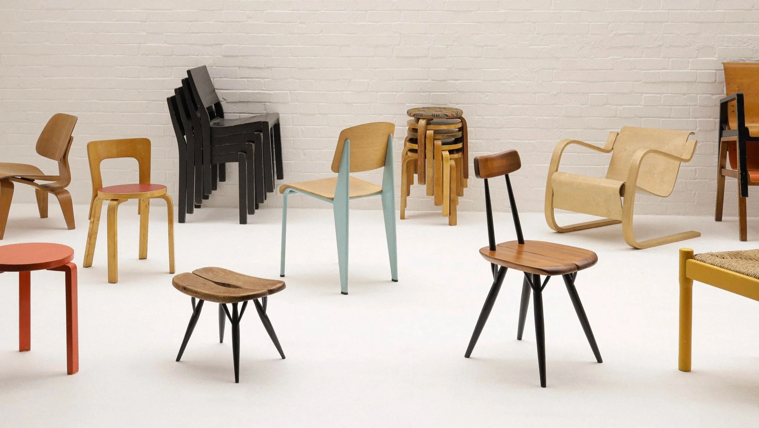 Collection of various iconic Finnish design chairs and stools arranged in a room with a white brick wall background. Featuring Alvar Aalto, Arne Jacobsen, Ilmari Tapiovaara, Jean Prouve, Vico Magistretti