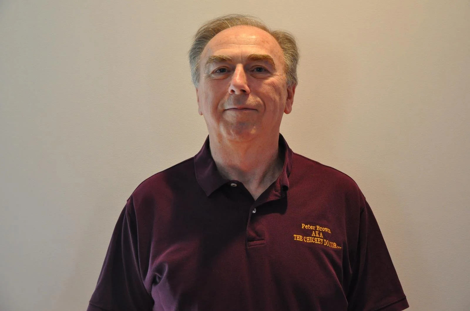 A middle-aged man with light hair and a slight smile standing against a plain beige wall. He is wearing a maroon polo shirt with yellow embroidered text that says "Peter Brown AKA THE CHICKEN DOCTOR..."