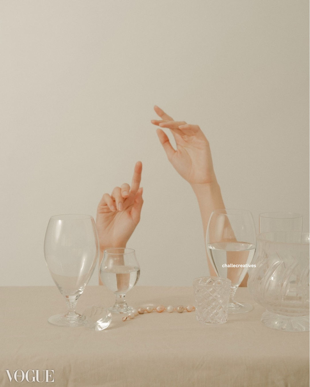 Two hands with extended fingers above a table with various glassware, including a water glass, a wine glass, and a textured small glass, arranged on a beige surface.