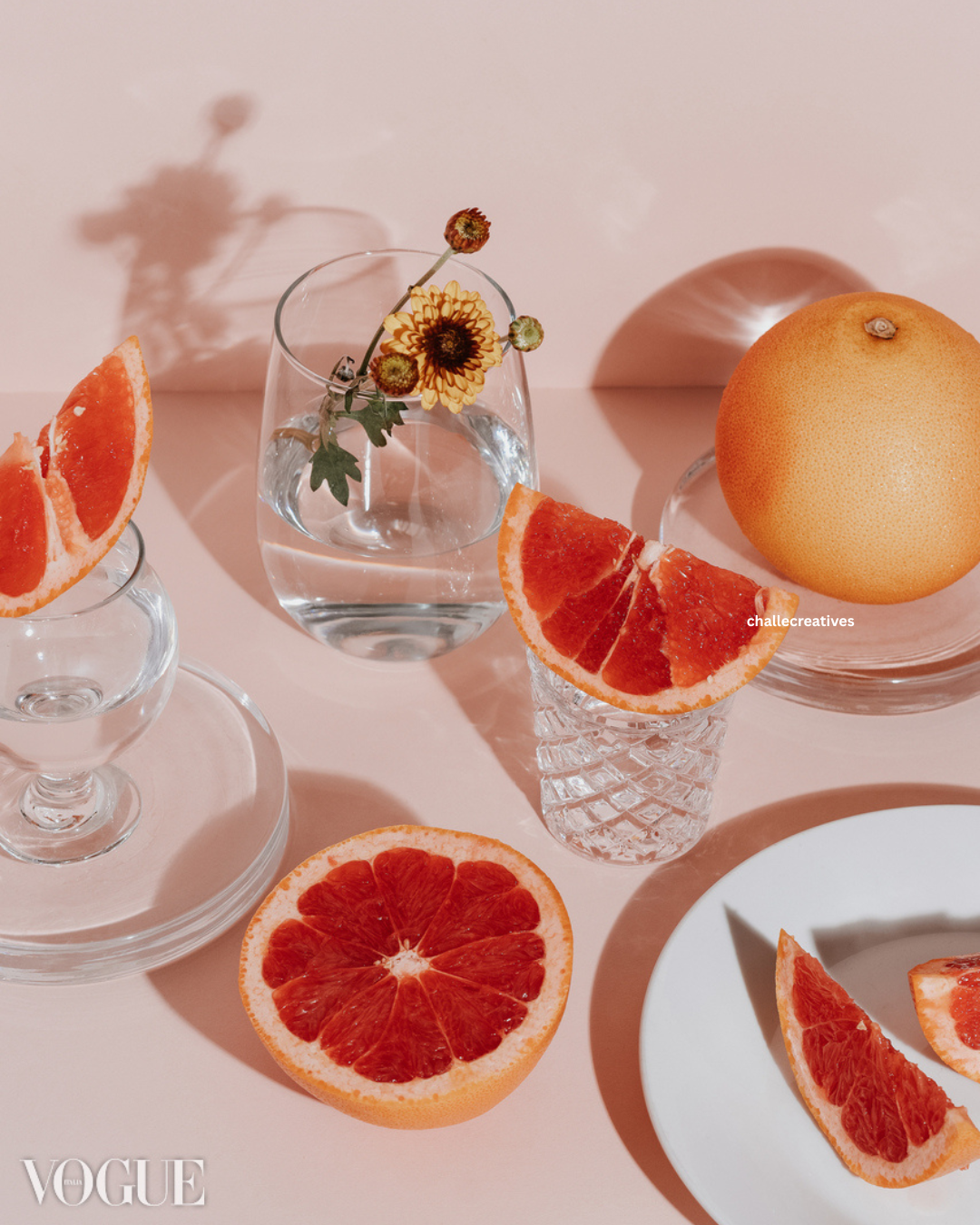 Decorative still life with grapefruit slices in glasses, a whole grapefruit on a glass plate, and a vase with a sunflower, on a pink surface with shadows.