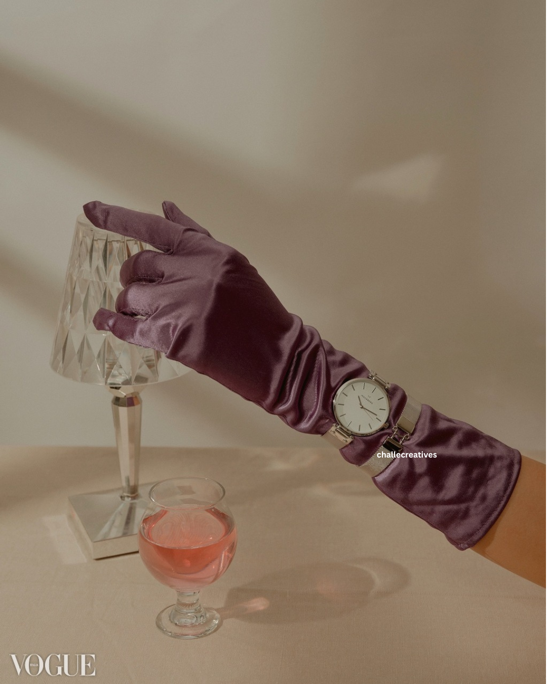 A person wearing a purple satin glove and a wristwatch is reaching to touch a table lamp with a diamond-patterned shade. A glass of pink liquid is on the table, and the VOGUE logo is in the bottom left corner.