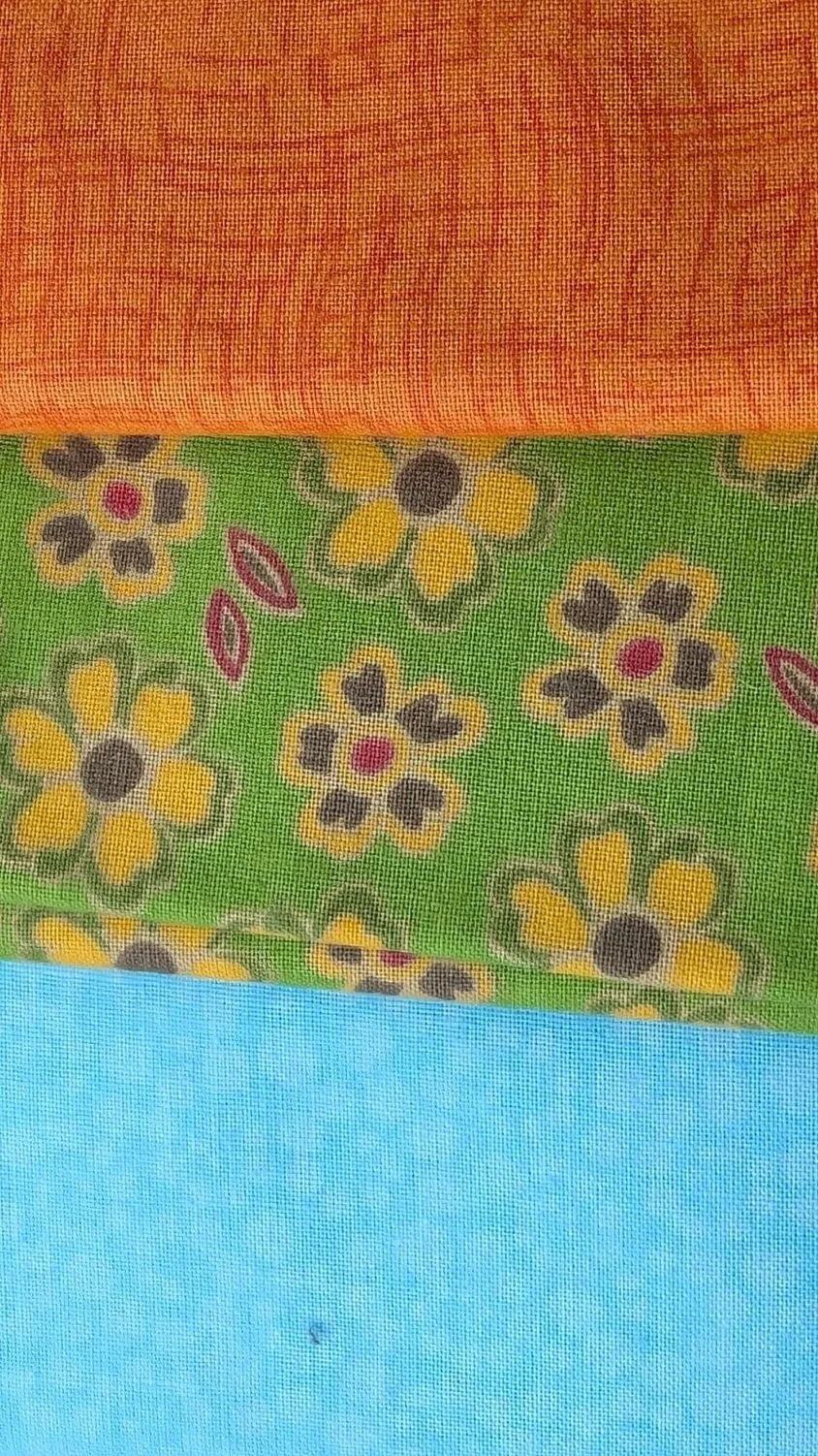 Close-up of fabric with three distinct patterns: a top orange textured pattern, a middle green floral pattern with yellow, black, and pink accents, and a bottom solid light blue fabric.