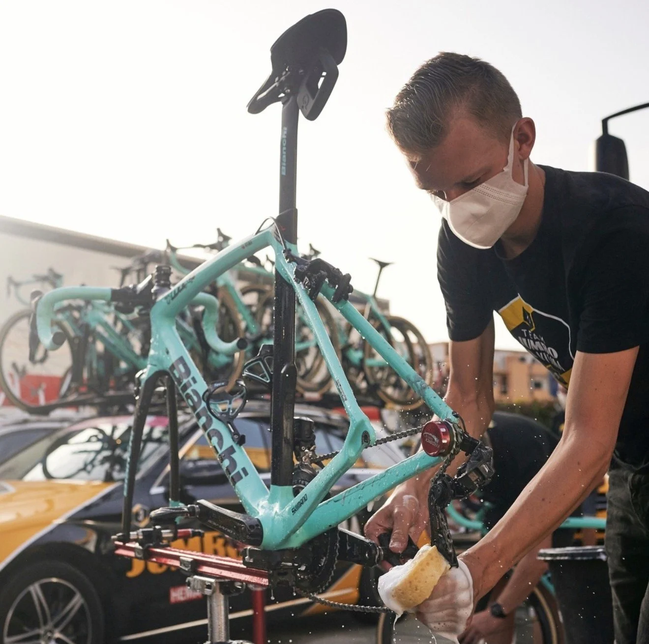 Persoon met mondkapje wast een racefiets met een spons bij een wielerwedstrijd of fietsevenement. Op de achtergrond staan meerdere fietsen op een bike stand en auto’s.