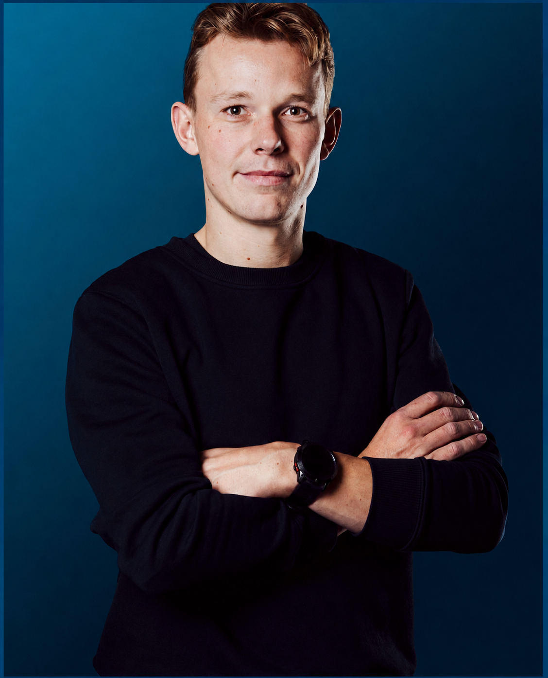 Portret van een jonge man met gekamd haar, zwarte trui en een smartwatch, met gekruiste armen, staand voor een blauwe achtergrond.