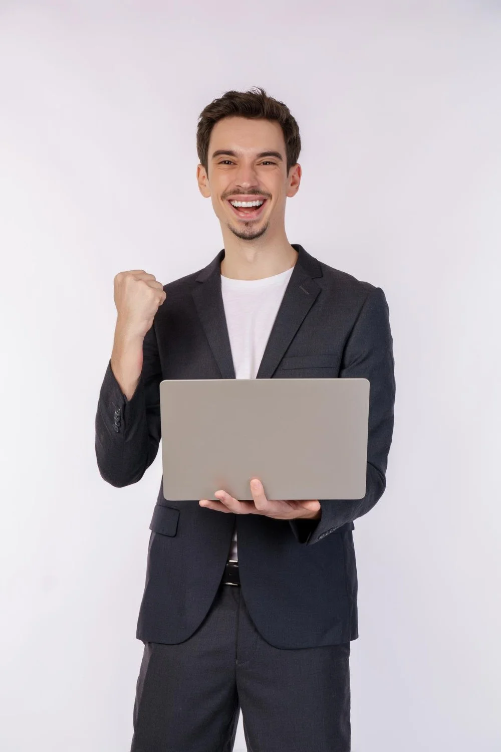 Young man in a business suit holding a laptop and smiling with a clenched fist gesture.