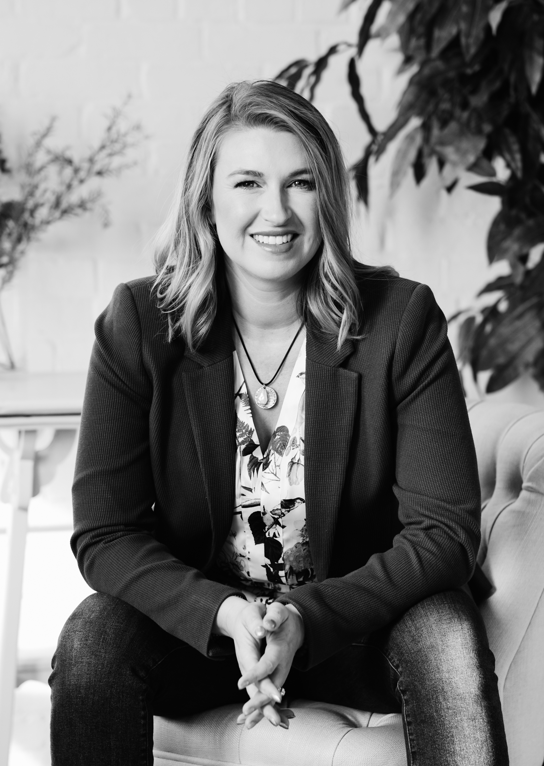 Black and white portrait of a woman with shoulder-length hair smiling, wearing a blazer over a floral blouse, sitting on a sofa with plants in the background.