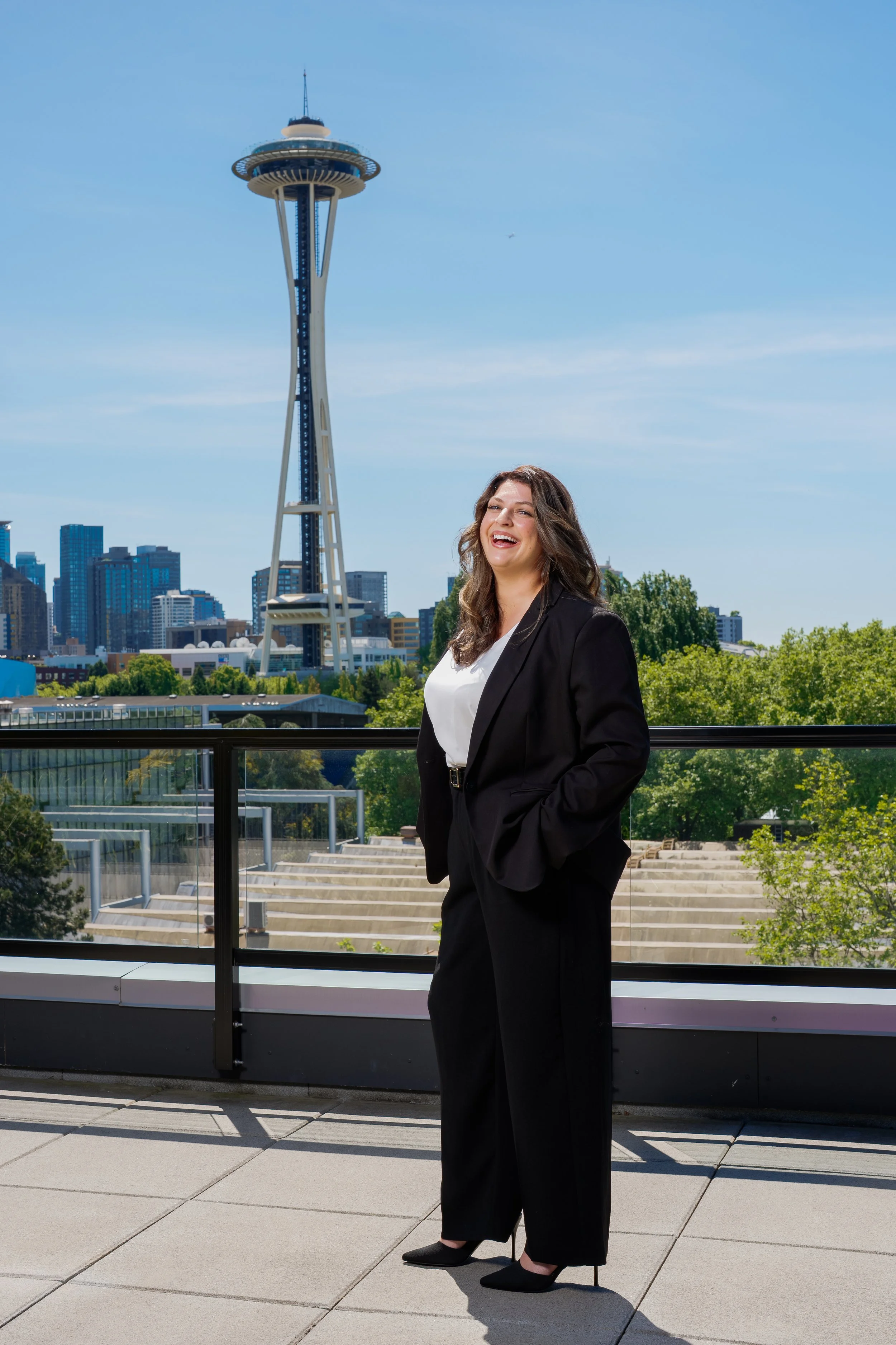 A woman in a black suit and high heels standing on a balcony with the Seattle skyline and Space Needle in the background during daytime.
