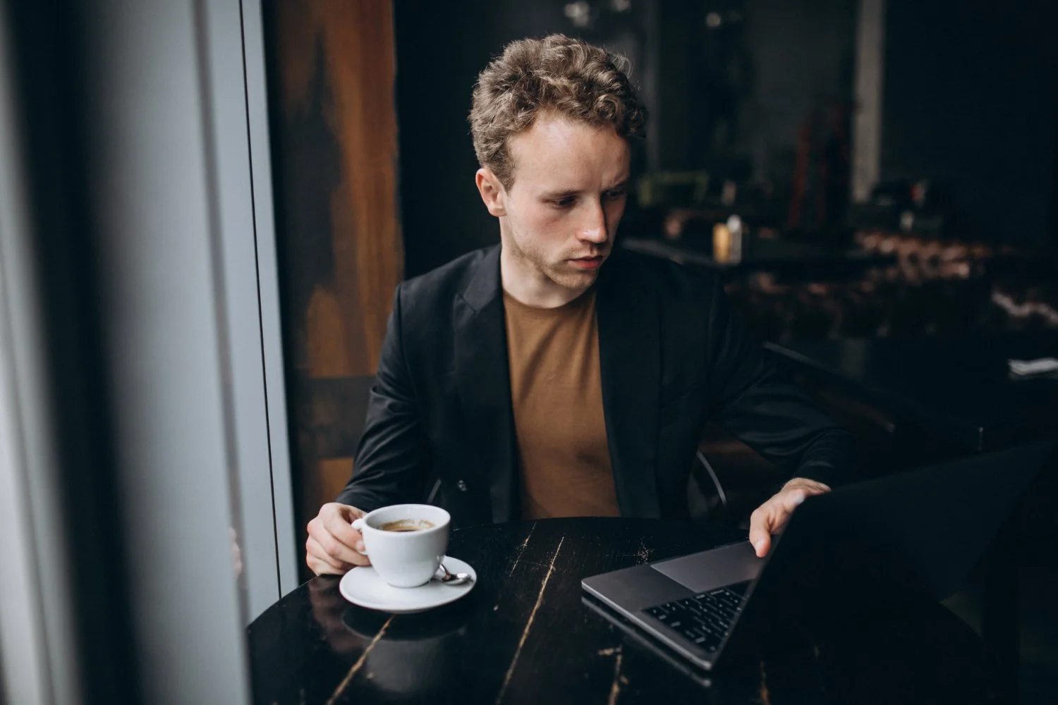 Young man with light brown curly hair working on a laptop in a dimly lit cafe while holding a cup of coffee.