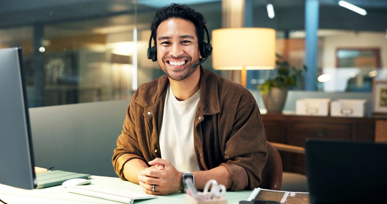Smiling man with dark curly hair wearing a headset sitting at a desk in a modern office.