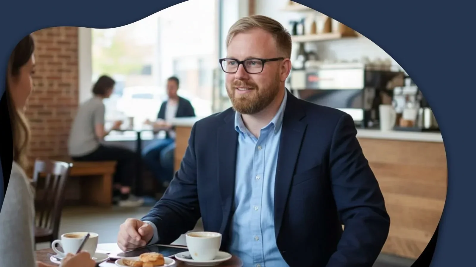 A man with glasses and a beard in a blue suit and light blue shirt, sitting at a table in a coffee shop, smiling and engaging in conversation with a woman. There are cups of coffee and a pastry on the table.