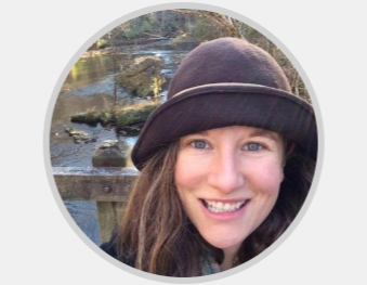 A smiling woman wearing a dark hat outdoors near a river with rocks and trees in the background.