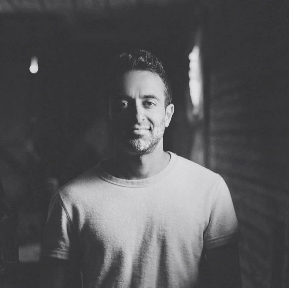 Black and white portrait of a smiling man with short hair and a beard, wearing a plain t-shirt, standing outdoors near a wooden wall.